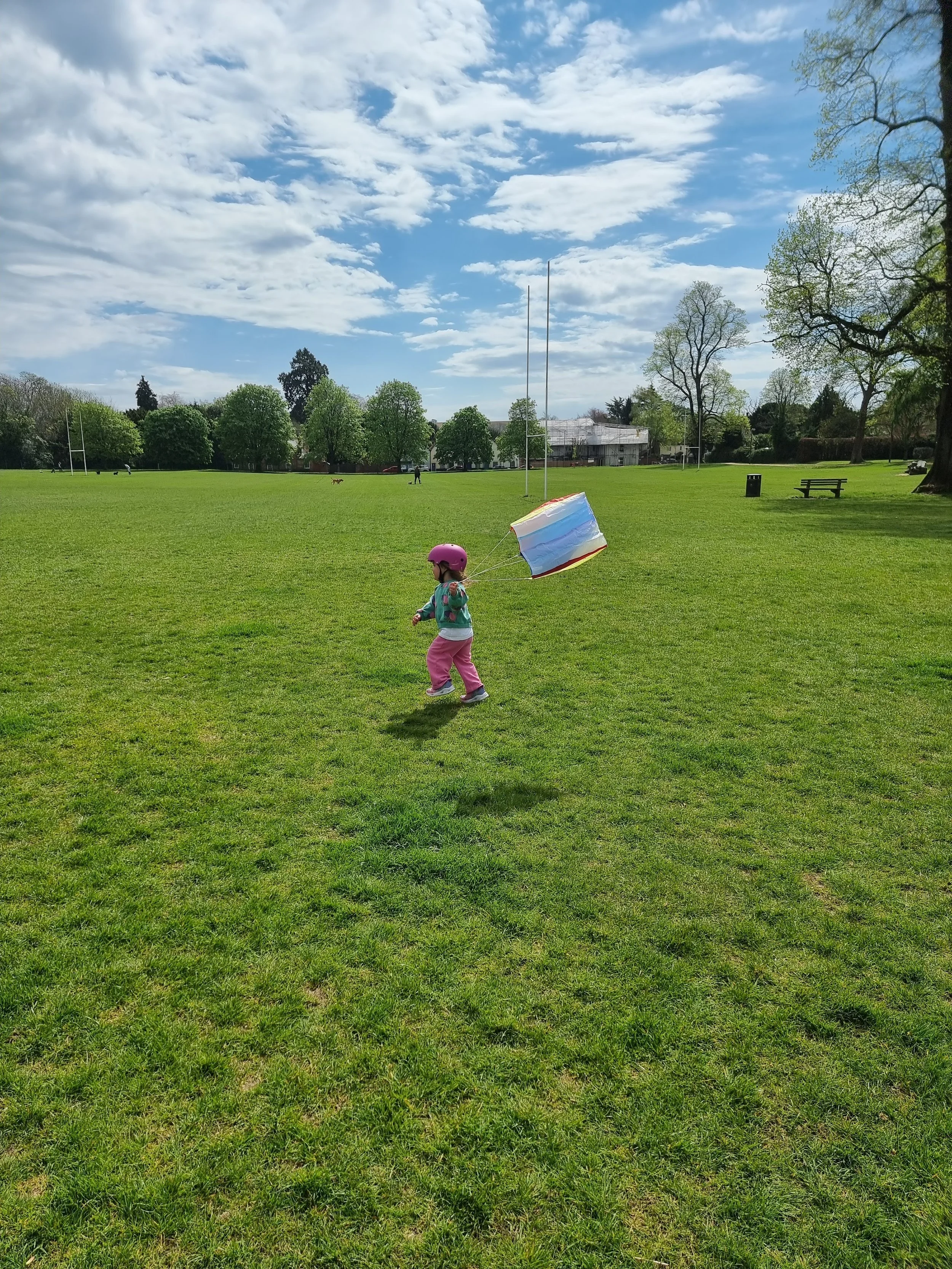 A young girl wearing a pink helmet, jacket, and pants runs on a grassy field while flying a small, multi-colored kite on a partly cloudy day.