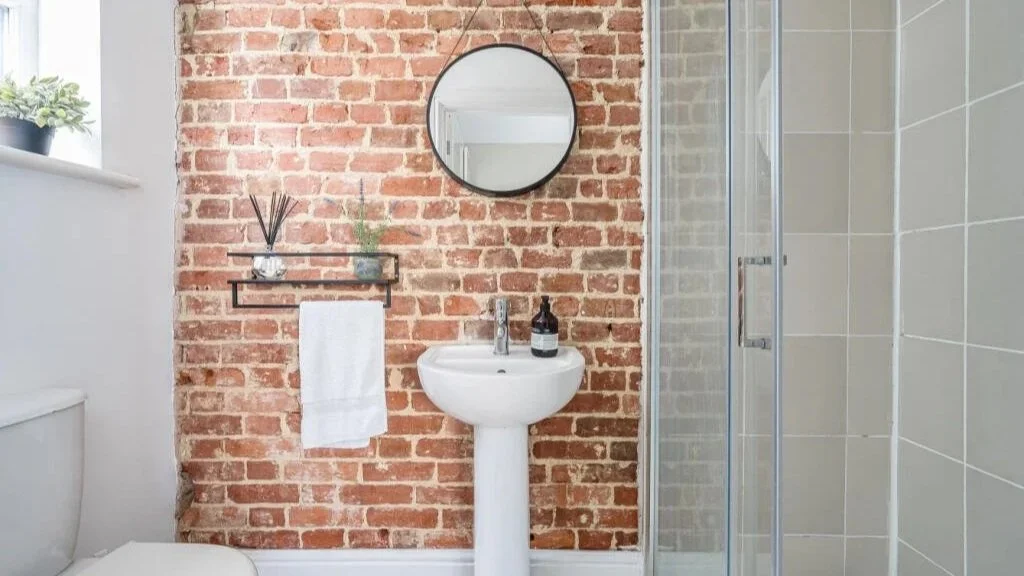 Small bathroom with a brick accent wall, white pedestal sink, round mirror, glass shower enclosure, white tiled wall, and a window with a potted plant.
