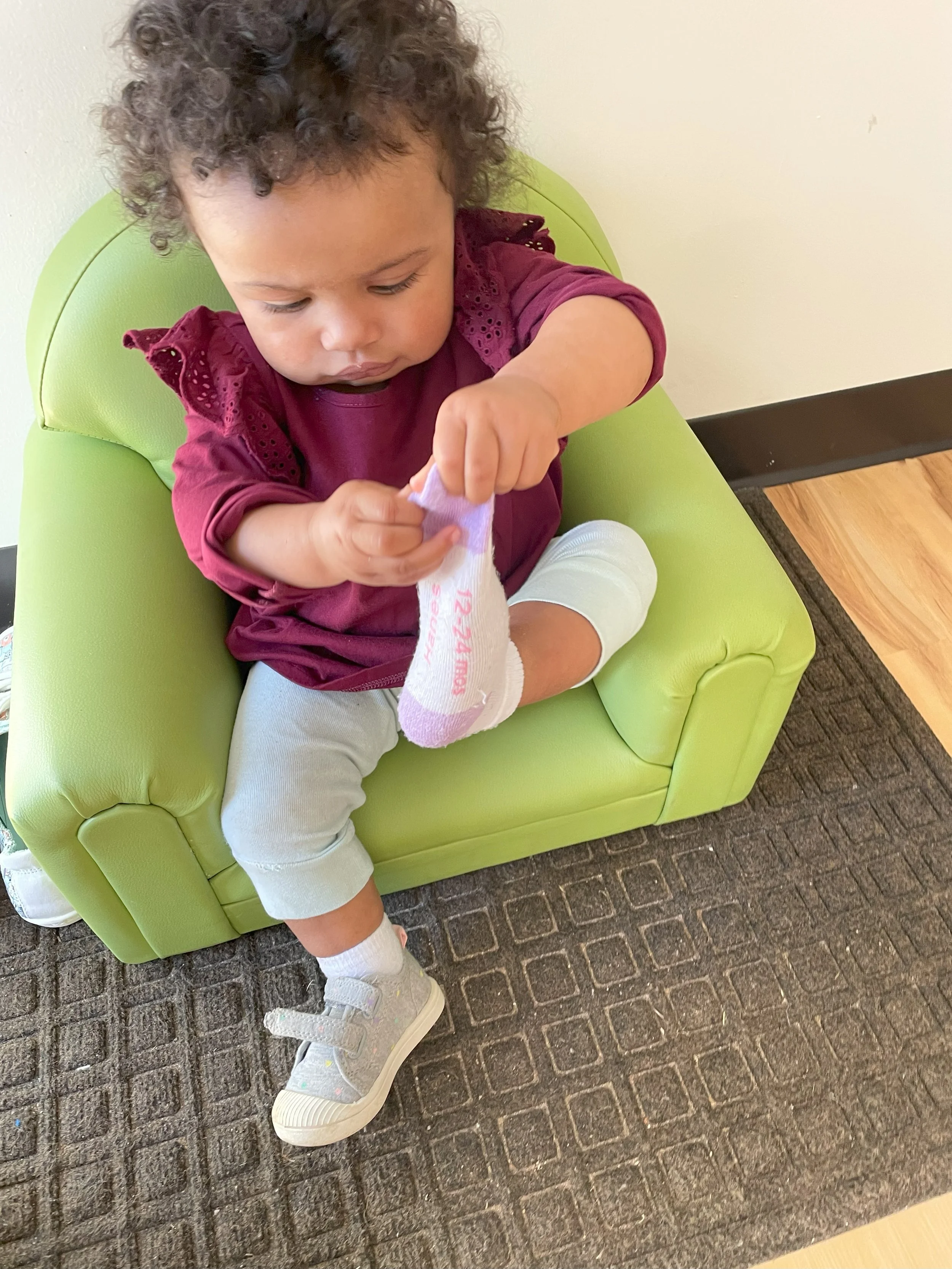 A young child sitting on a green chair, putting on a purple sock while wearing white shoes and light-colored pants.