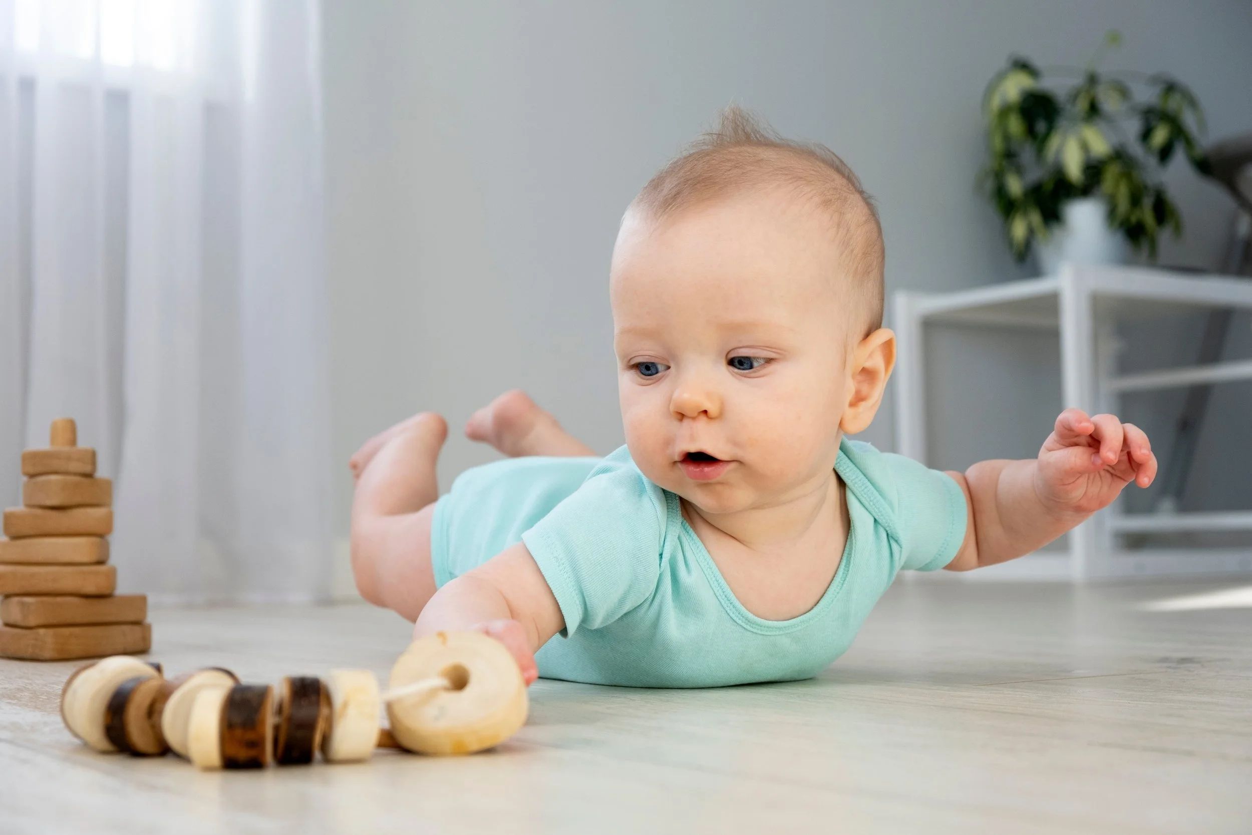 A baby in a mint green shirt lying on a wooden floor, playing with wooden beads and a ring toy.