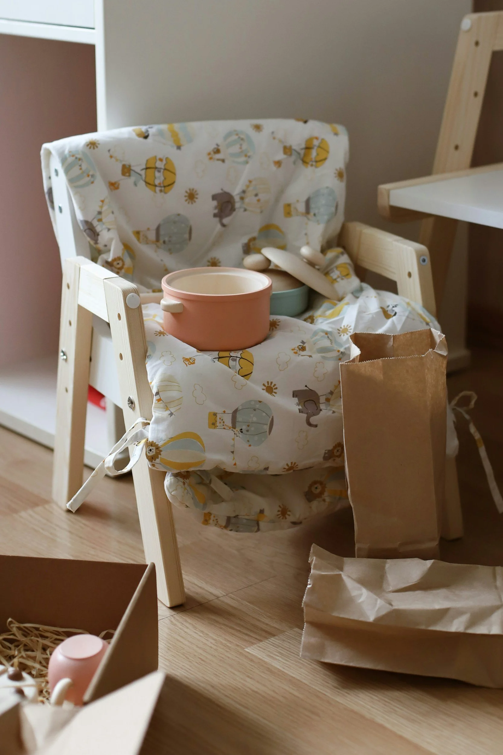 A child’s wooden high chair with a colorful animal-themed cushion, placed near a white shelf. There are pink and light green bowls on the chair, along with a wooden spoon. A brown paper bag and a crumpled paper bag are on the floor nearby.