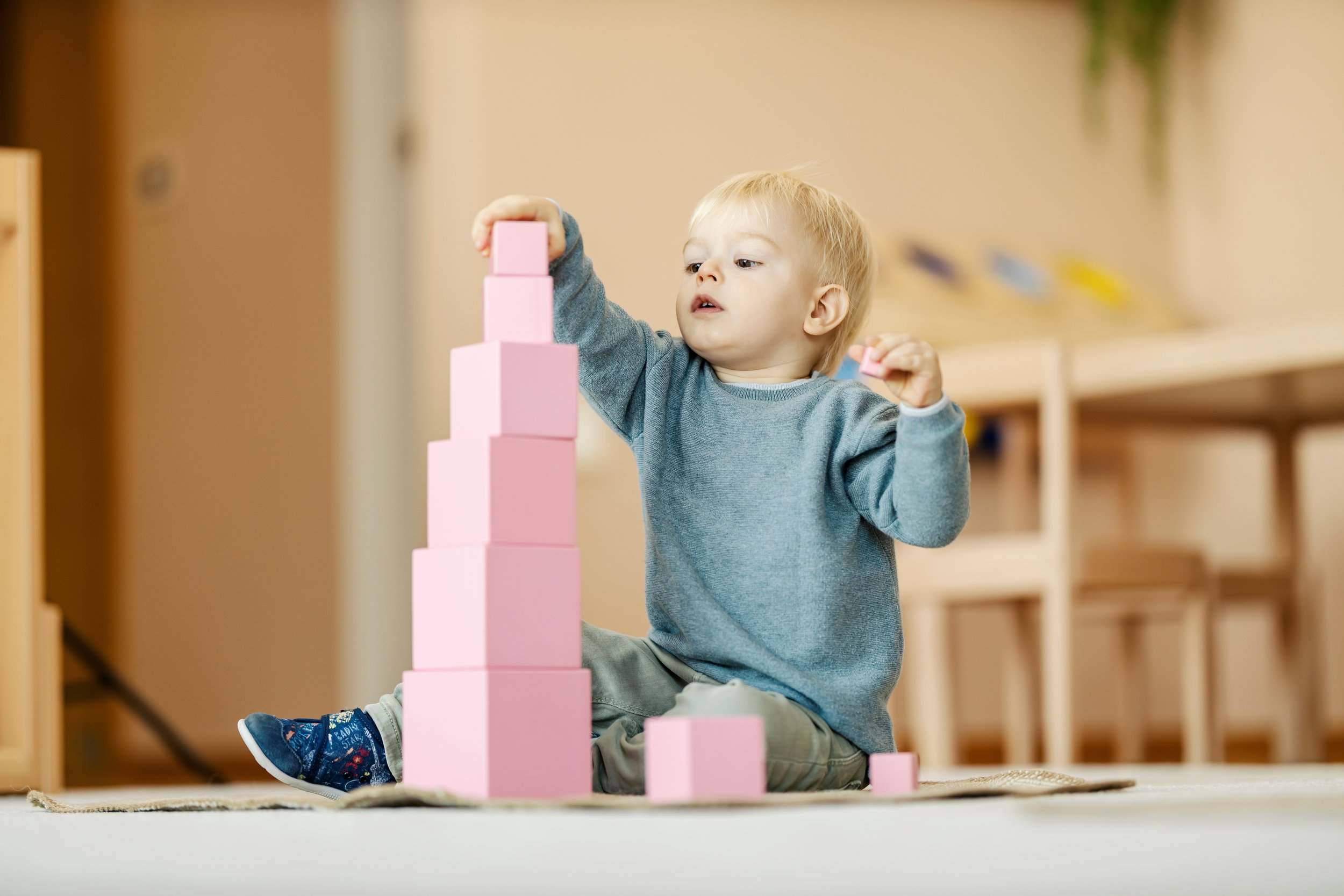 A young boy in a blue sweater and gray pants playing with pink foam blocks on the floor of a room.