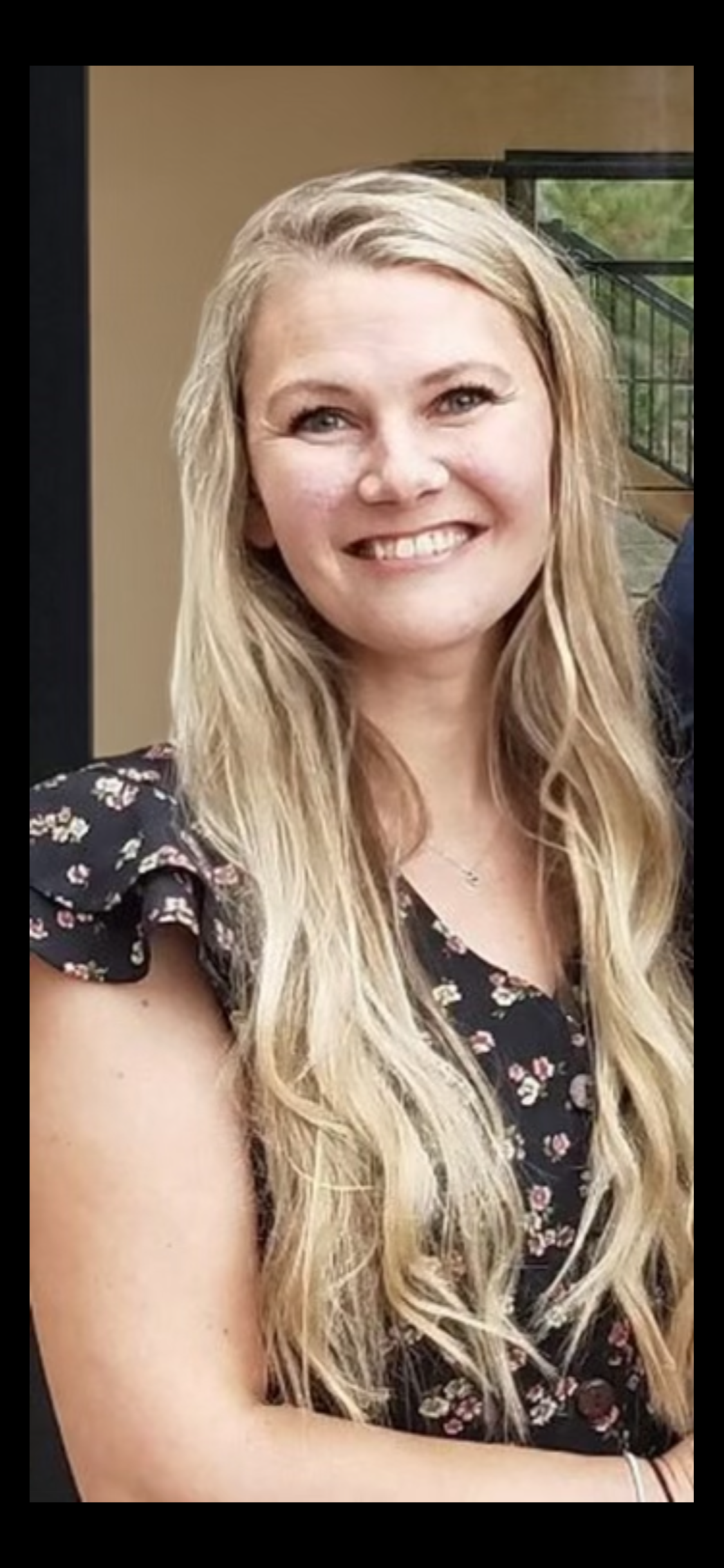 A woman with long blonde hair smiling, wearing a black floral dress with ruffled sleeves, standing indoors near a window with a black frame.