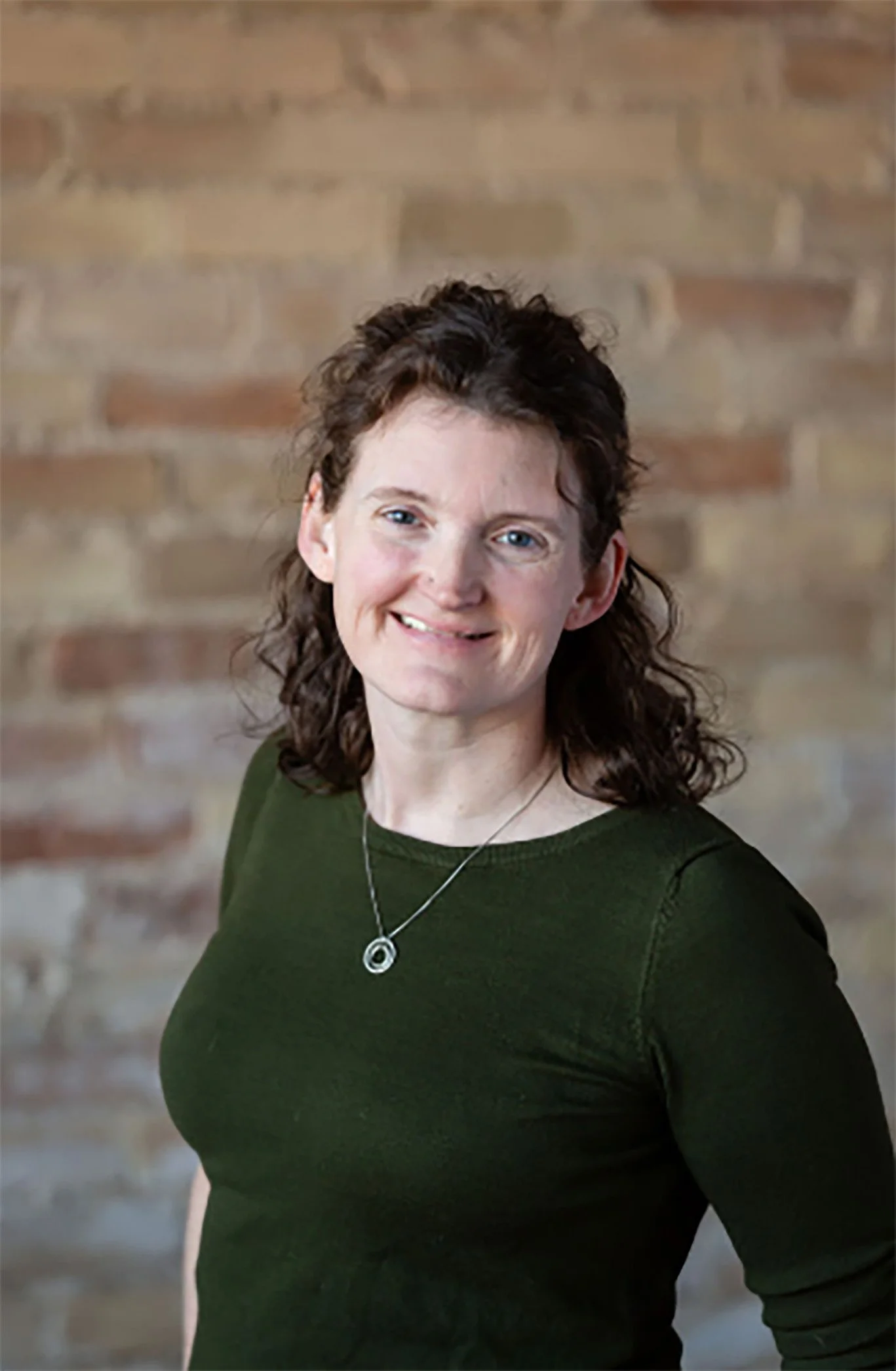 A woman with curly brown hair and blue eyes smiling at the camera, wearing a dark green shirt and a silver necklace, standing in front of a brick wall.