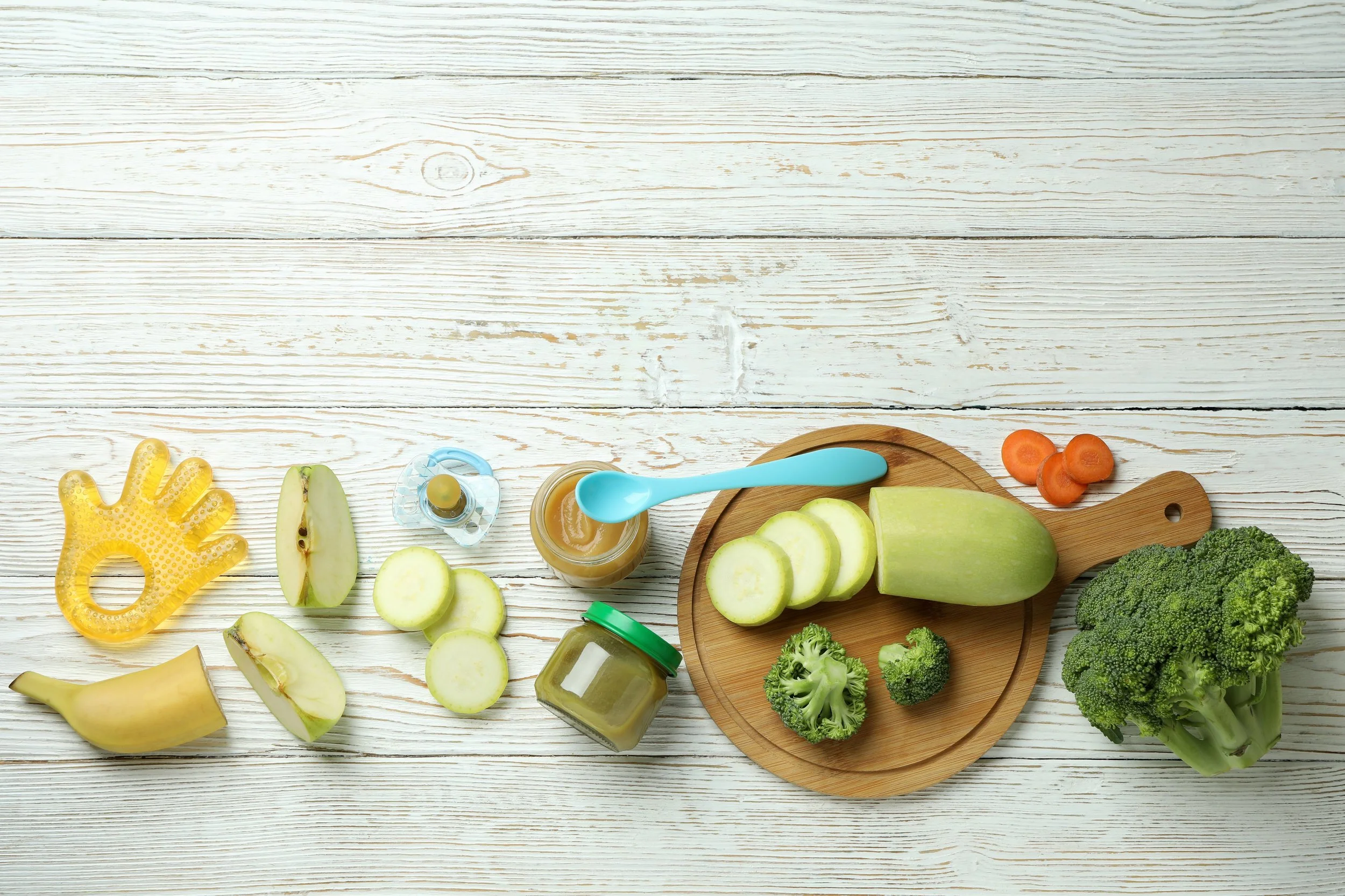 Assorted vegetables, fruit slices, a jar of green sauce, baby food jars, and a blue spoon on a white wooden surface.