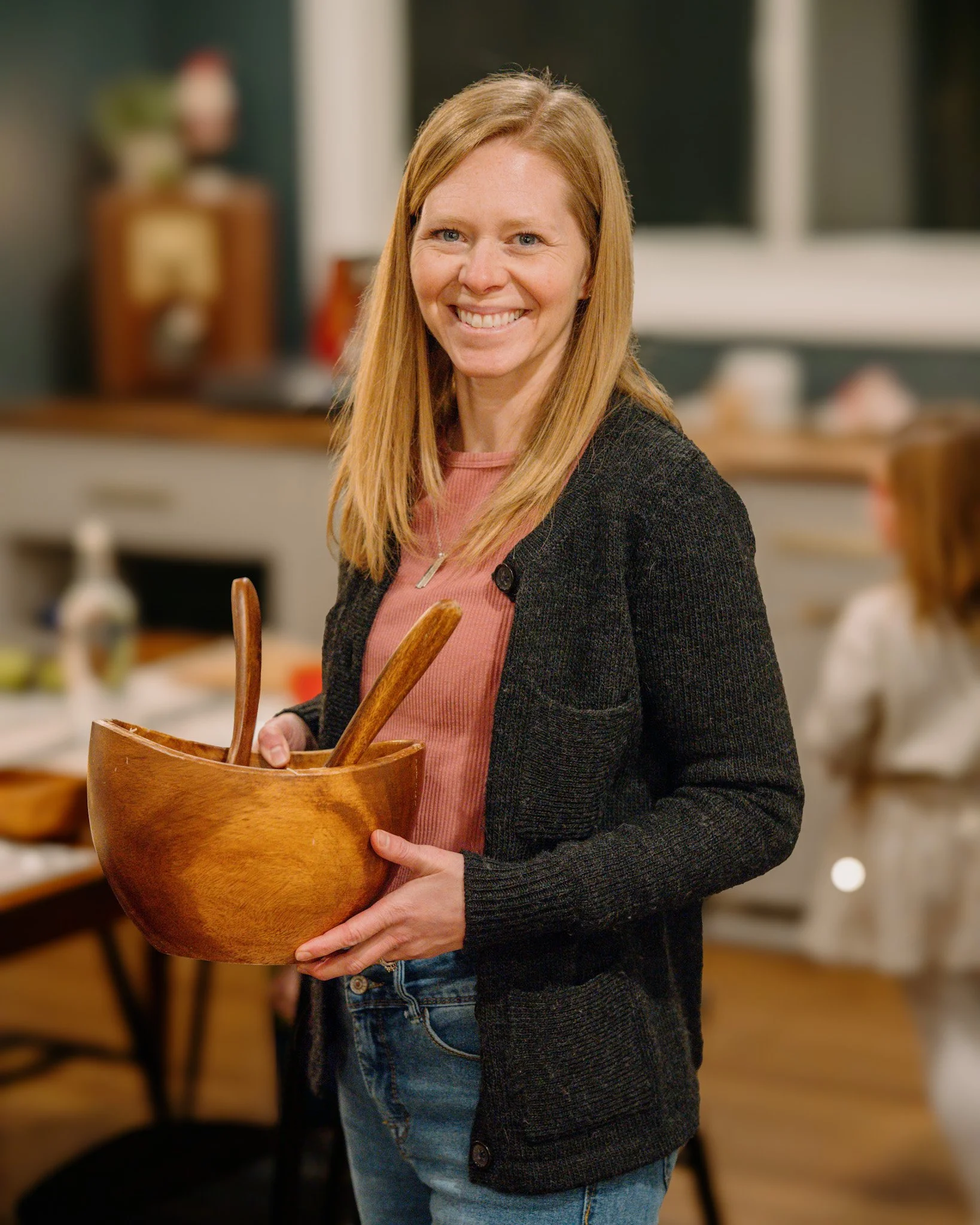A woman with red hair smiling and holding a wooden mixing bowl with kitchen utensils inside, in a cozy home kitchen.