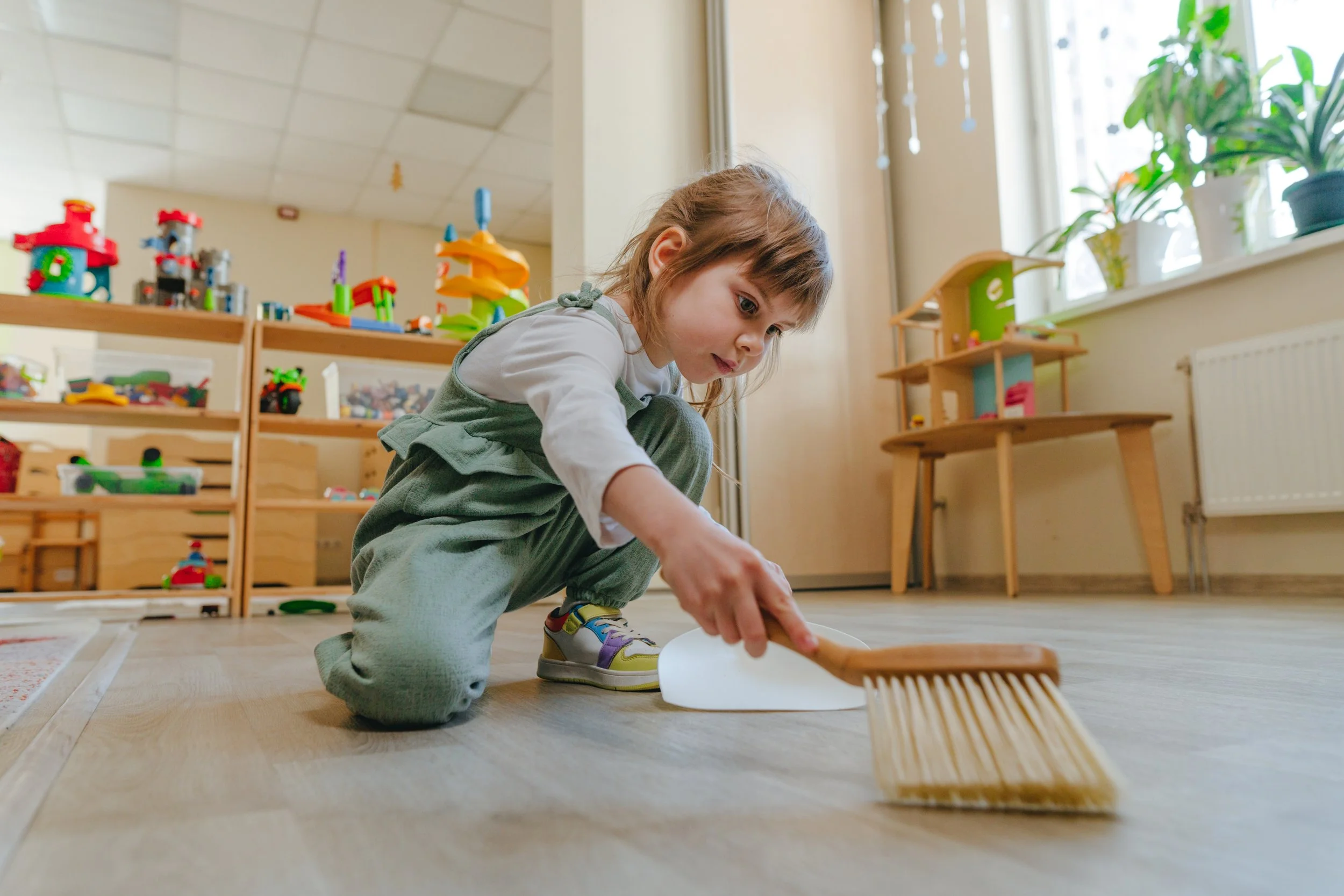 A young girl crouches on the floor sweeping with a small broom in a playroom filled with toys and potted plants.