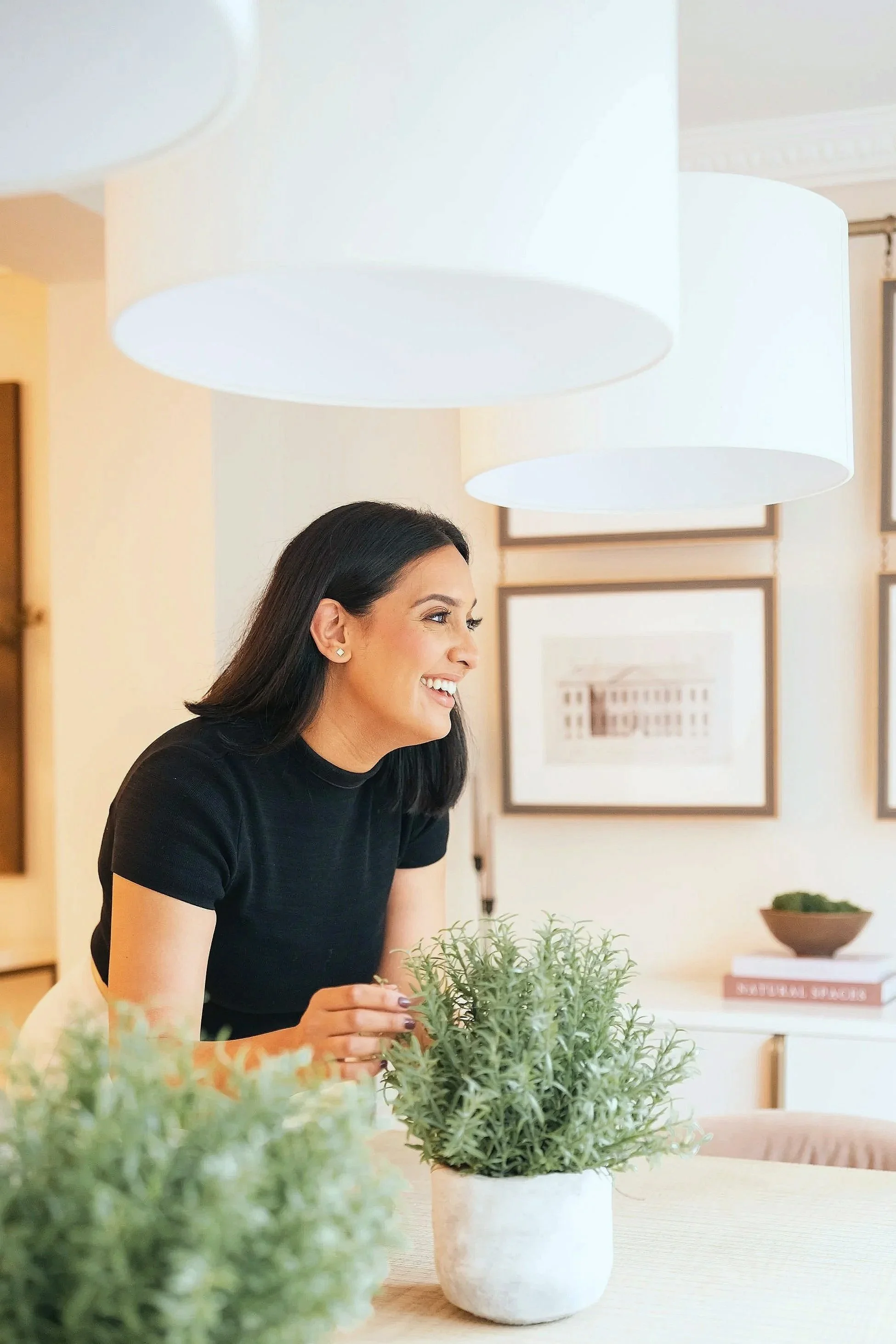 A woman smiling, sitting at a table with potted plants in a well-lit, modern room with framed artwork on the wall.