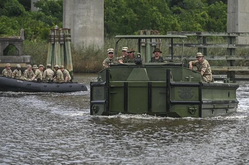 A group of soldiers in military uniform riding on a military amphibious vehicle on a river, with some soldiers in a black inflatable boat behind them.