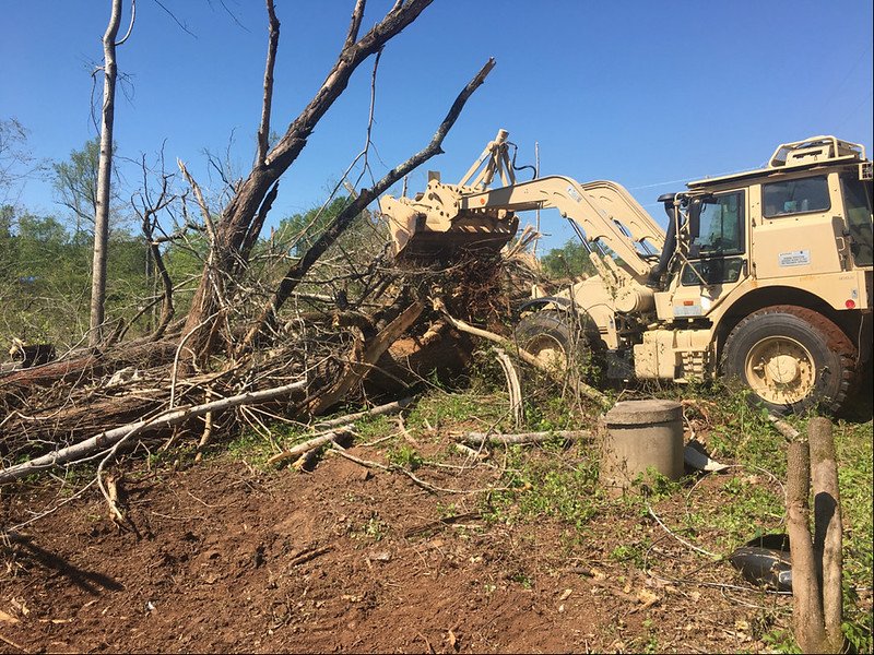 Excavator clearing fallen trees and debris in a forested area under a clear blue sky.
