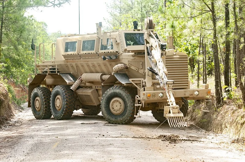A large military vehicle, possibly an armored personnel carrier, blocking a dirt road in a wooded area.