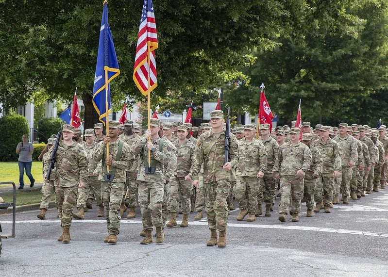 Group of soldiers marching in uniform during a parade, carrying flags including the American flag, with trees and spectators in background.