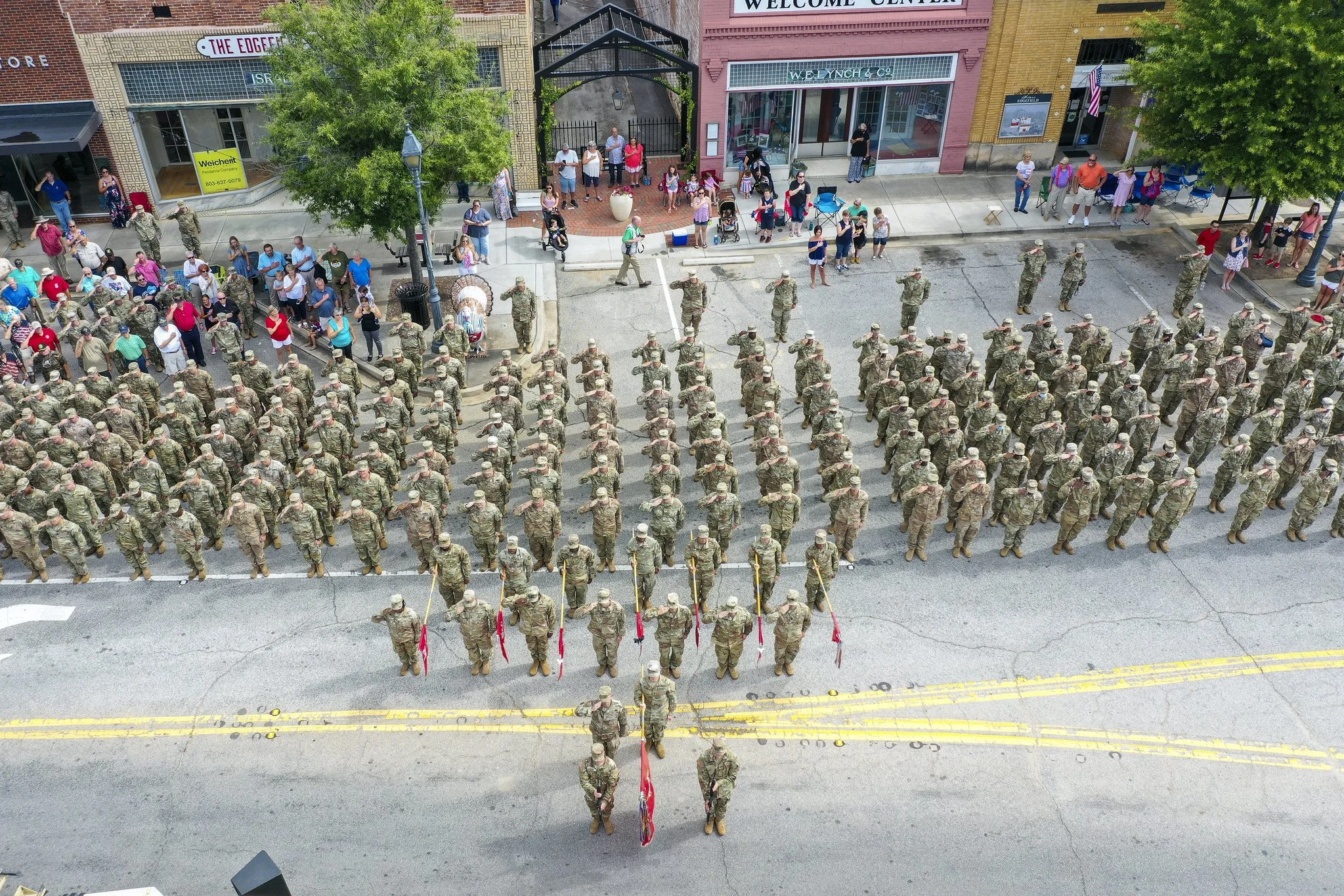 Aerial view of a large group of soldiers in uniform standing in formation on a city street during a parade, with spectators and storefronts lining the sidewalk.