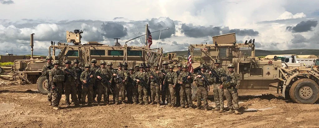 Group of soldiers standing in front of military vehicles on a dirt field under cloudy sky.