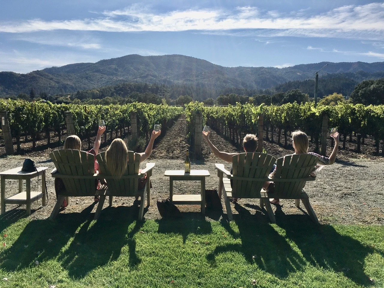 Four people sitting on lounge chairs outdoors, raising wine glasses, with a vineyard and mountains in the background.