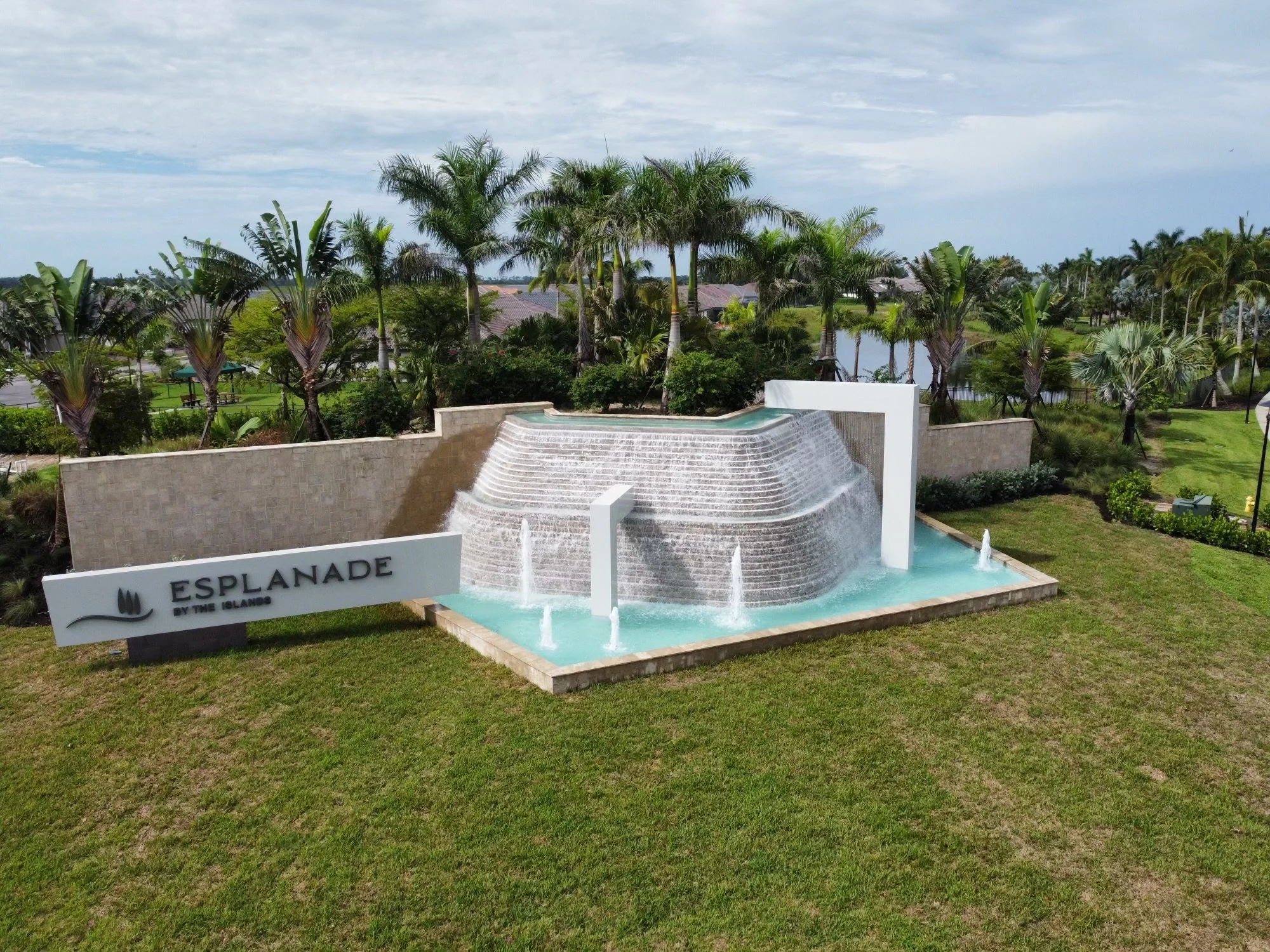 A decorative water feature with cascading waterfalls and fountains, situated outdoors with lush green grass, palm trees, and a body of water in the background, and a sign that reads 'Esplanade by the Islands.'