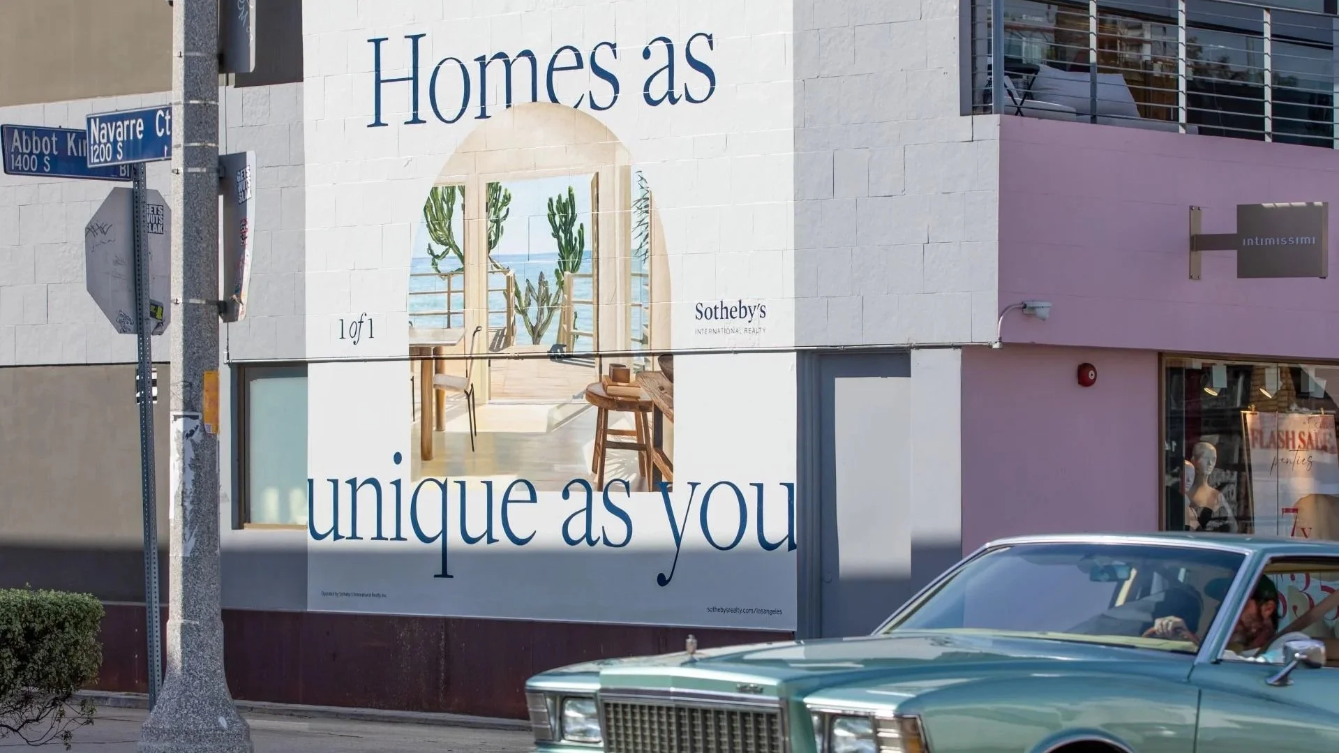 Street view of a building with an advertisement mural promoting homes as unique as you, featuring interior decor and potted cacti, with street signs for Abbot Kinney and Navarre Court, and a vintage car driving past.