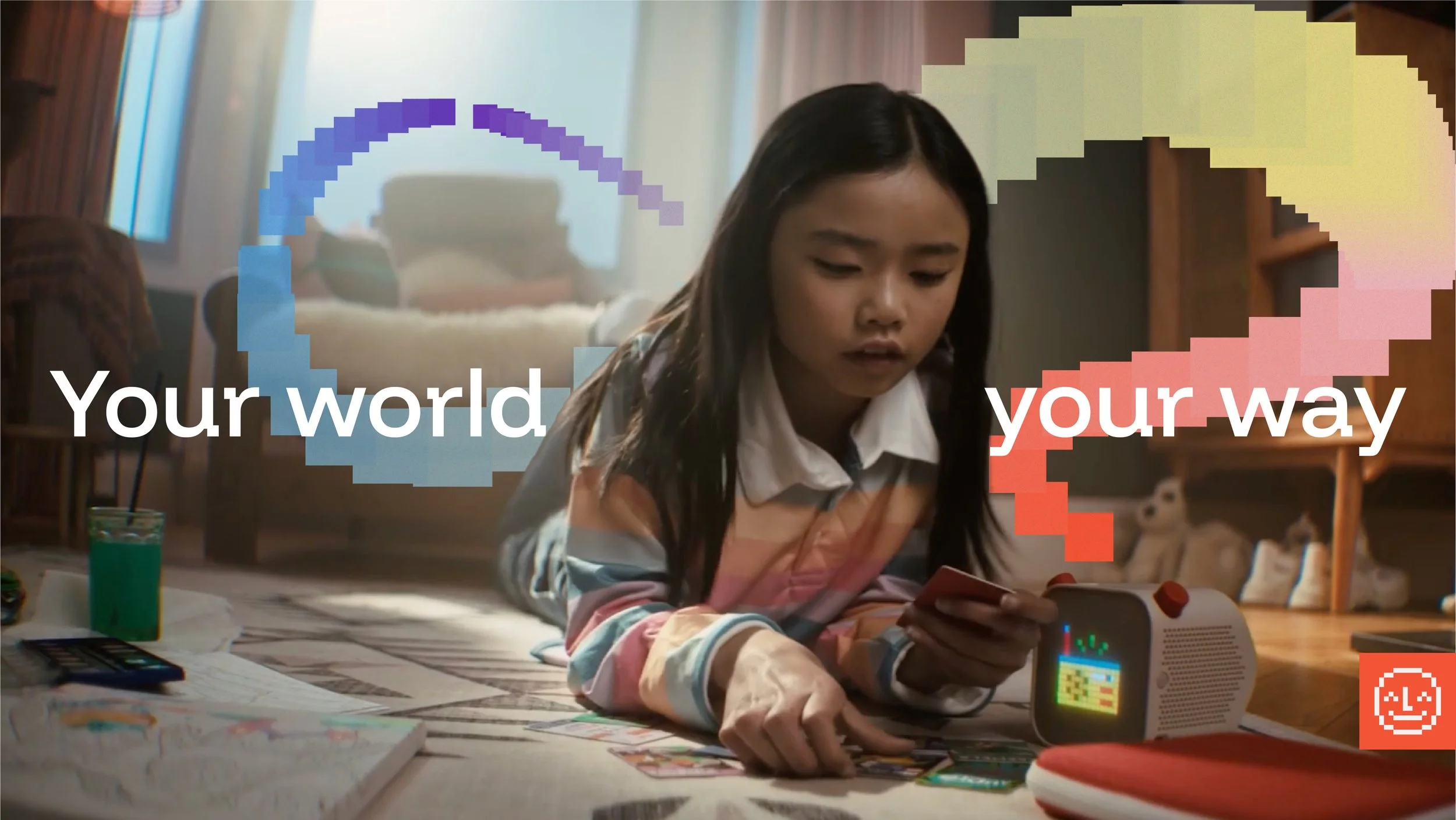 A young girl lying on her stomach on the floor, looking at her phone with a small electronic device displaying a colorful pixelated image nearby. The background shows a cozy room with a bed, stuffed animals, and various toys and books on the floor. O