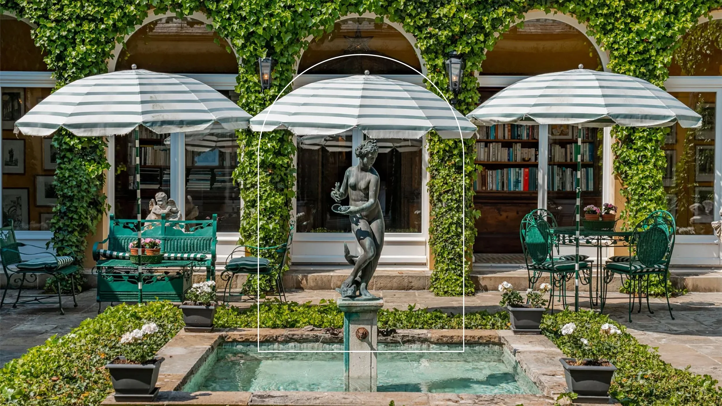 A garden patio with a fountain featuring a statue of a woman holding a bowl, surrounded by potted flowers, green bushes, a round table with four chairs, two large striped umbrellas, and a house with green vine-covered arches and large windows in the 