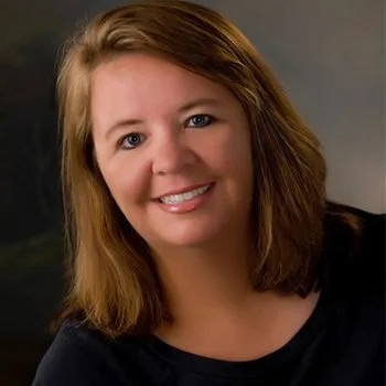 A woman with shoulder-length light brown hair and blue eyes smiling at the camera, wearing a black top against a dark background.