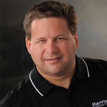 Portrait of a man in a black polo shirt with white trim, smiling against a blurred background.