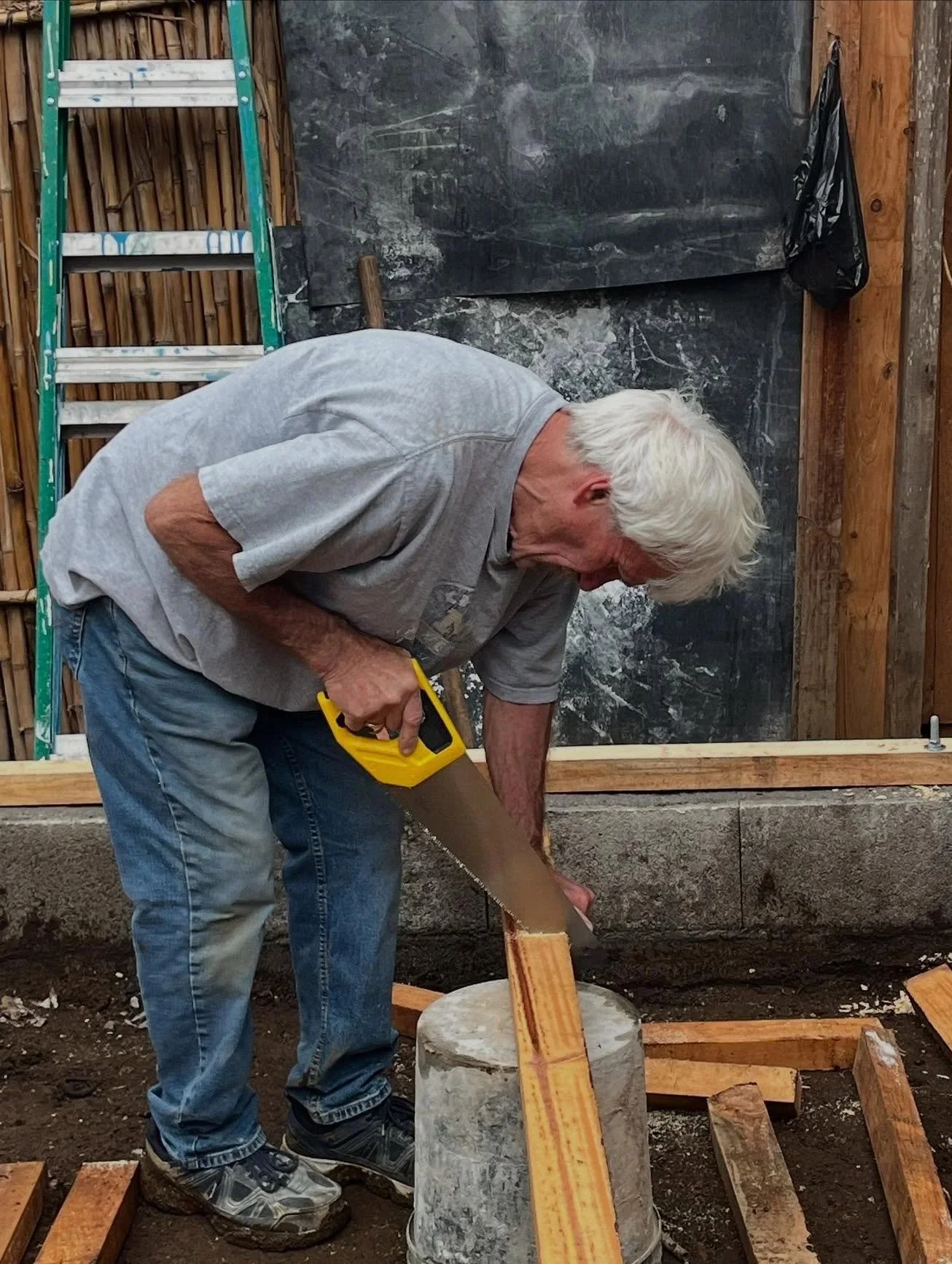 An elderly man with white hair working on a construction site, sawing a piece of wood with a hand saw, while standing on a concrete block. There is a ladder and wooden framing in the background.