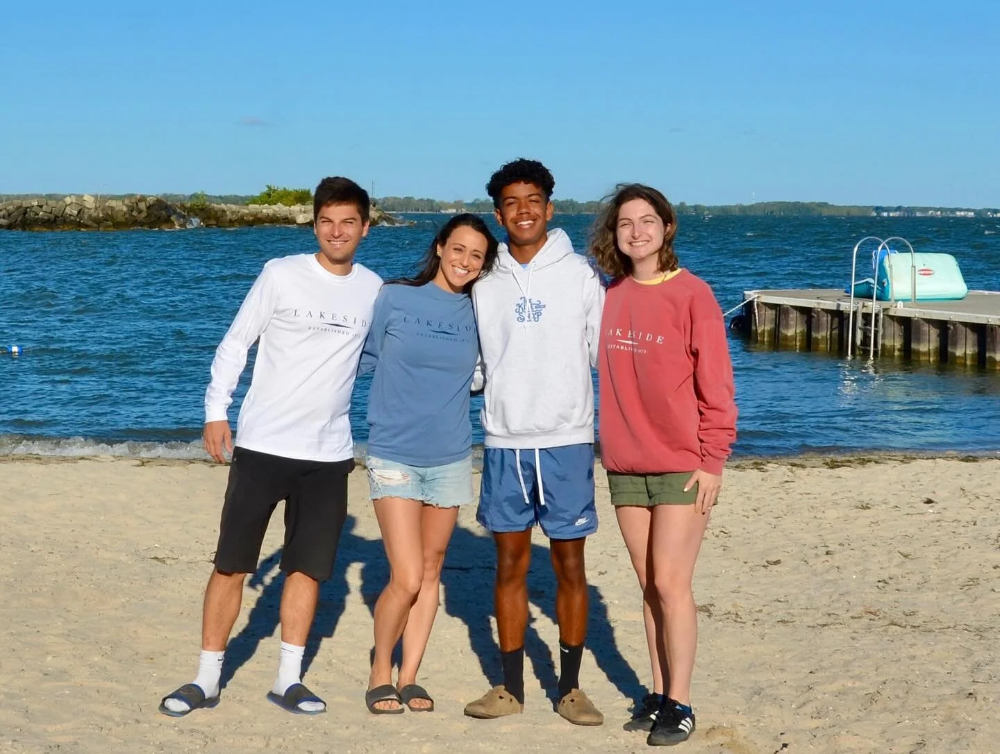 Four friends standing on a sandy beach near the water, posing together and smiling. They are dressed casually with some wearing matching 'Lakeside' sweatshirts. The background shows a shoreline, water, and a small dock with a ladder.