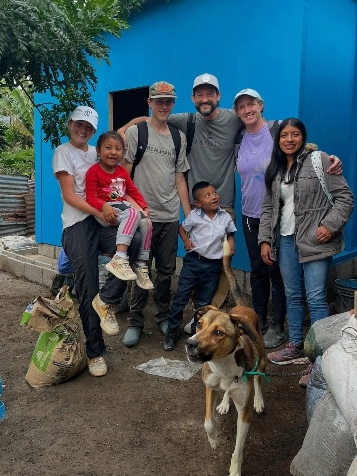 Group of seven people, including children and adults, posing outdoors with a dog in front of a blue building. Some are smiling, and one child is holding a fish.