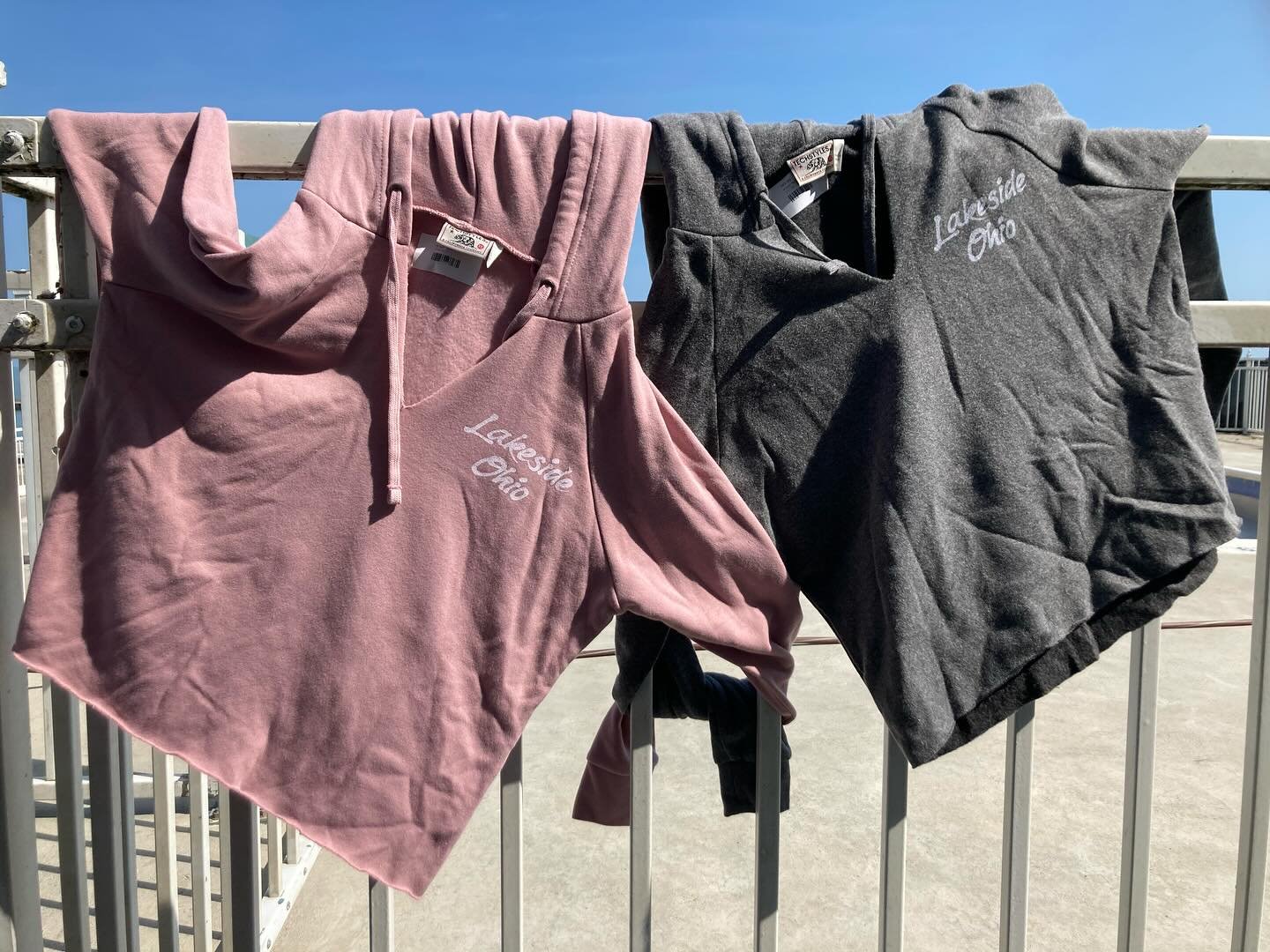 Two velour sweatshirts, one pink and one dark gray, hanging on a metal railing outside under a clear blue sky. Both sweatshirts have 'Lakeside Ohio' embroidered on the left chest area.
