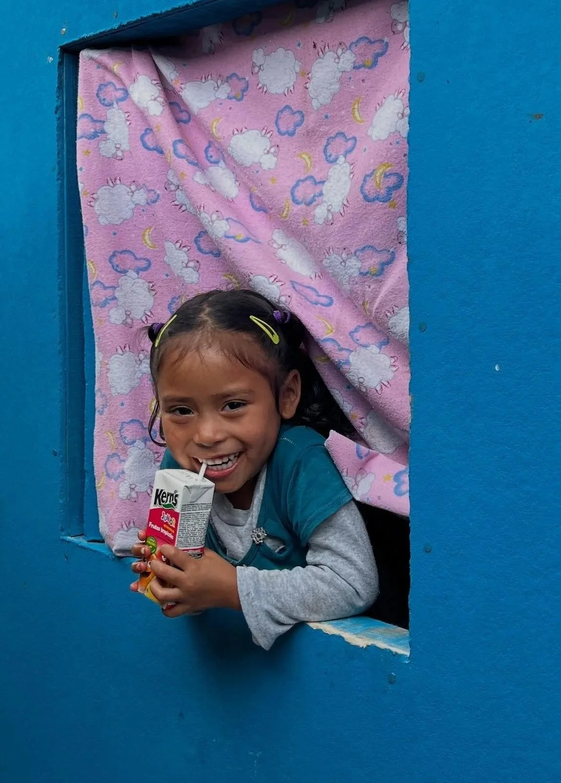 A young girl smiling and drinking from a juice box while leaning out of a small window with a pink curtain with clouds and moons, set in a blue wall.