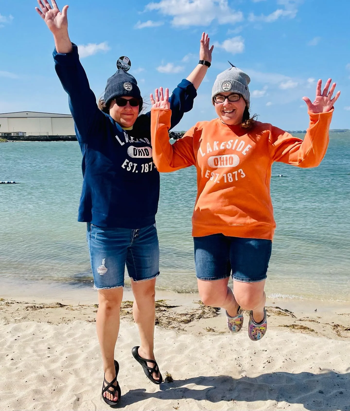 Two women jumping on a sandy beach near the water, wearing hoodies with 'Lakeside Ohio Est. 1873' printed on them, with one in a blue hoodie and the other in an orange hoodie, both wearing gray beanies and shorts, smiling and celebrating.
