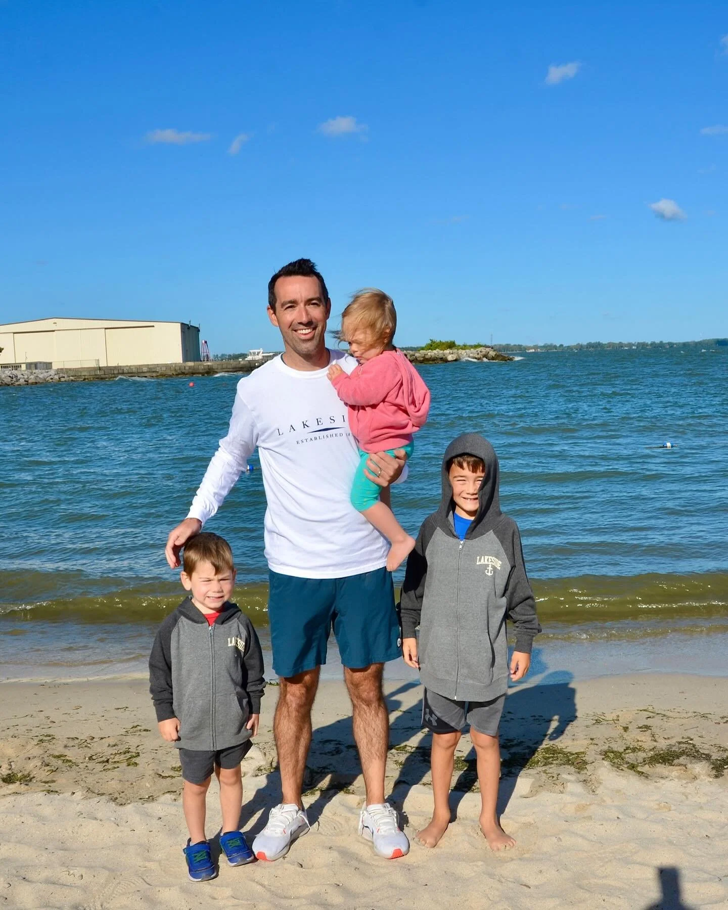 A man and three children standing on a beach near the water, smiling at the camera. The man is holding a girl in a pink hoodie, and two boys in gray hoodies are standing beside him.
