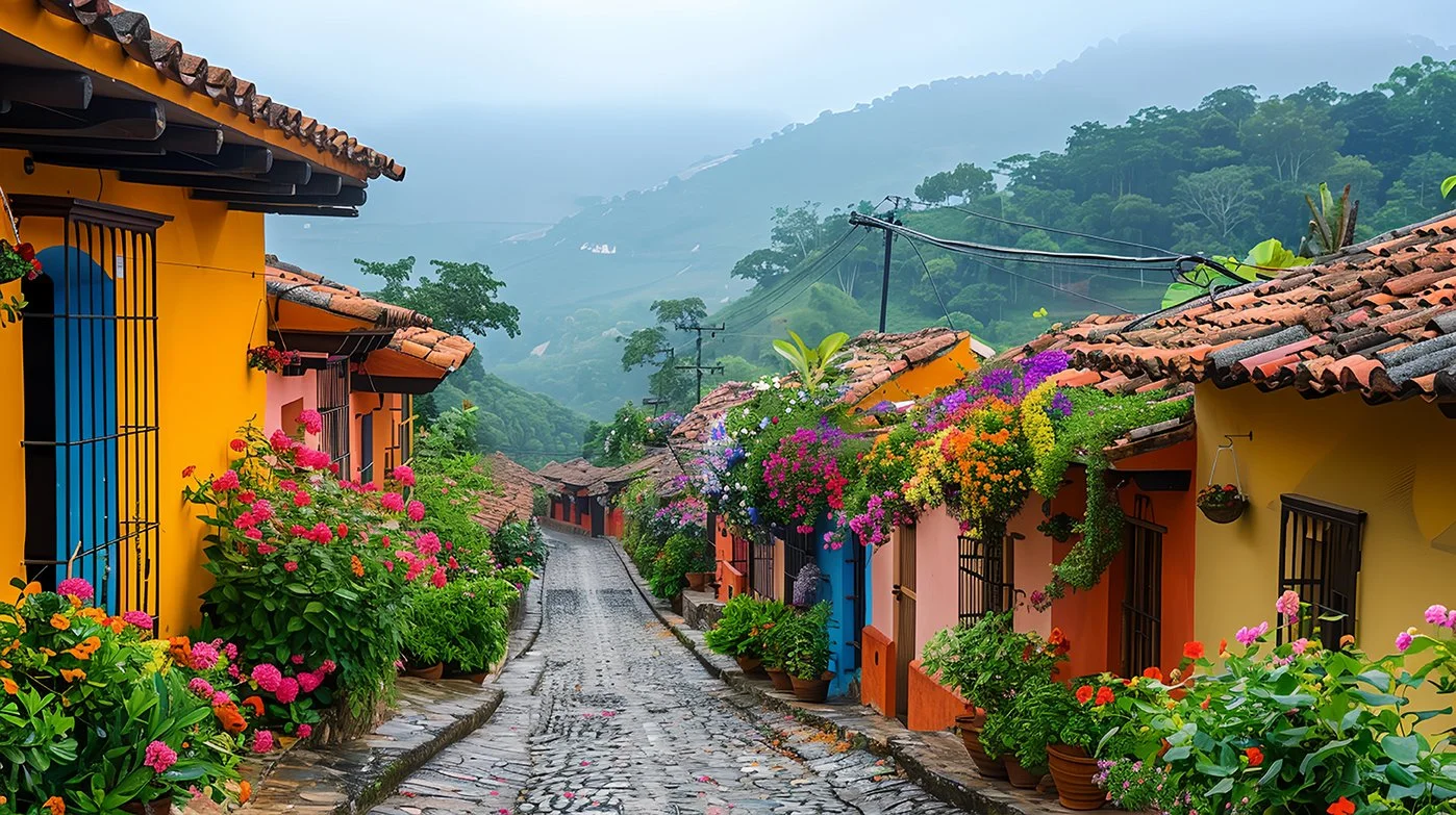 Colorful houses with red-tiled roofs along a cobblestone street, decorated with vibrant flowers in pots, situated in a lush, mountainous landscape.