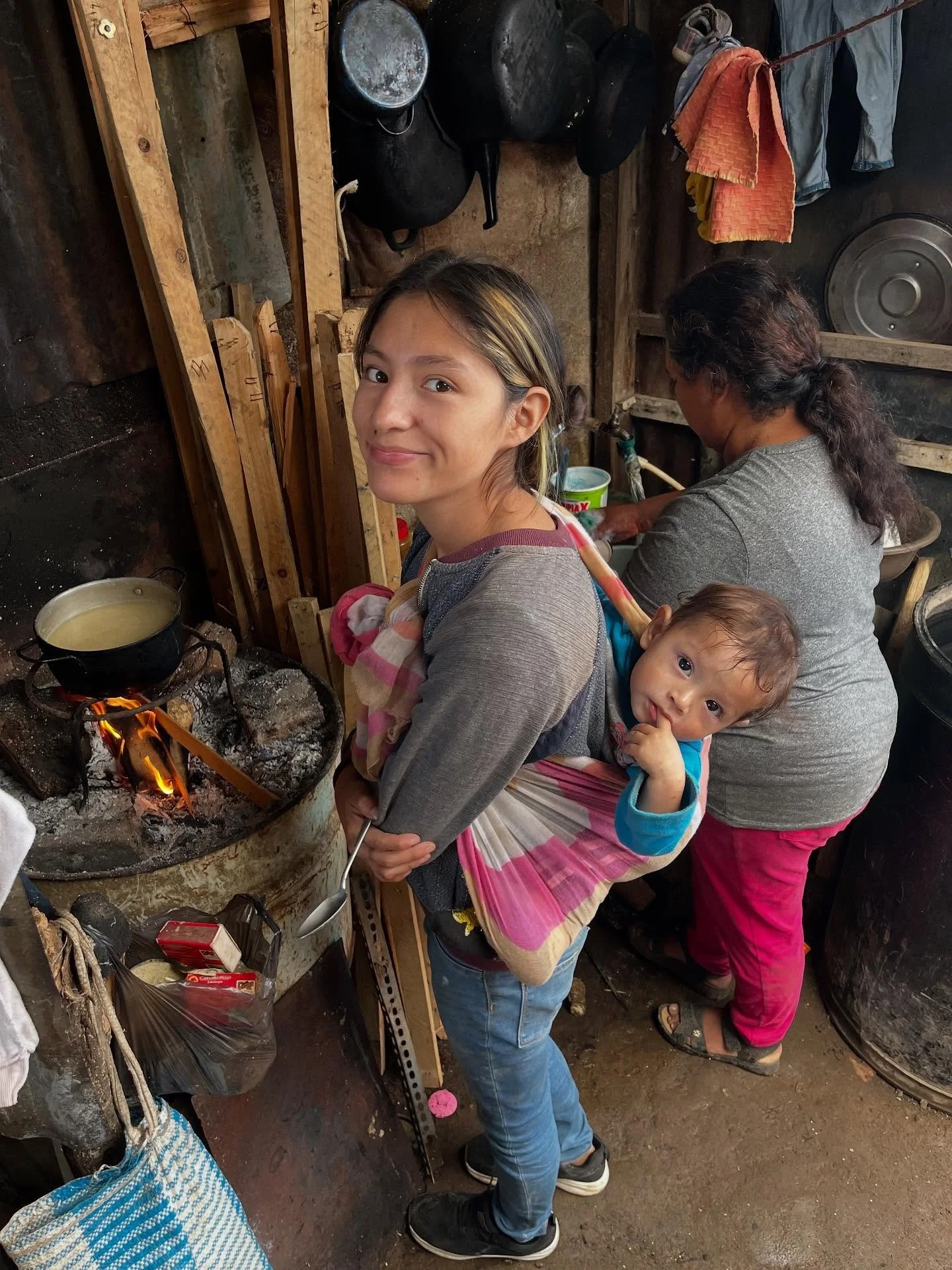 A young girl with blonde hair is carrying a toddler on her back using a colorful wrap. They are in a rustic kitchen with a wood-fueled stove, and a woman is cooking in the background. The kitchen appears simple and functional, with hanging pots and utensils.
