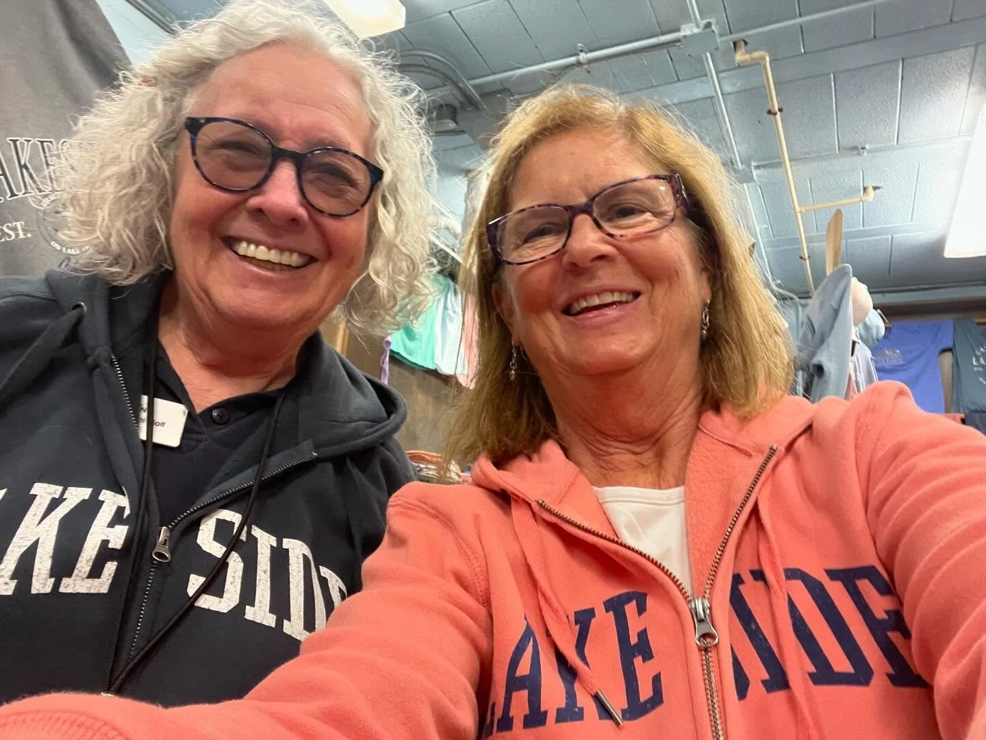 Two women smiling and taking a selfie in a room with merchandise and clothing in the background, both wearing 'Fake Sid' branded hoodies.