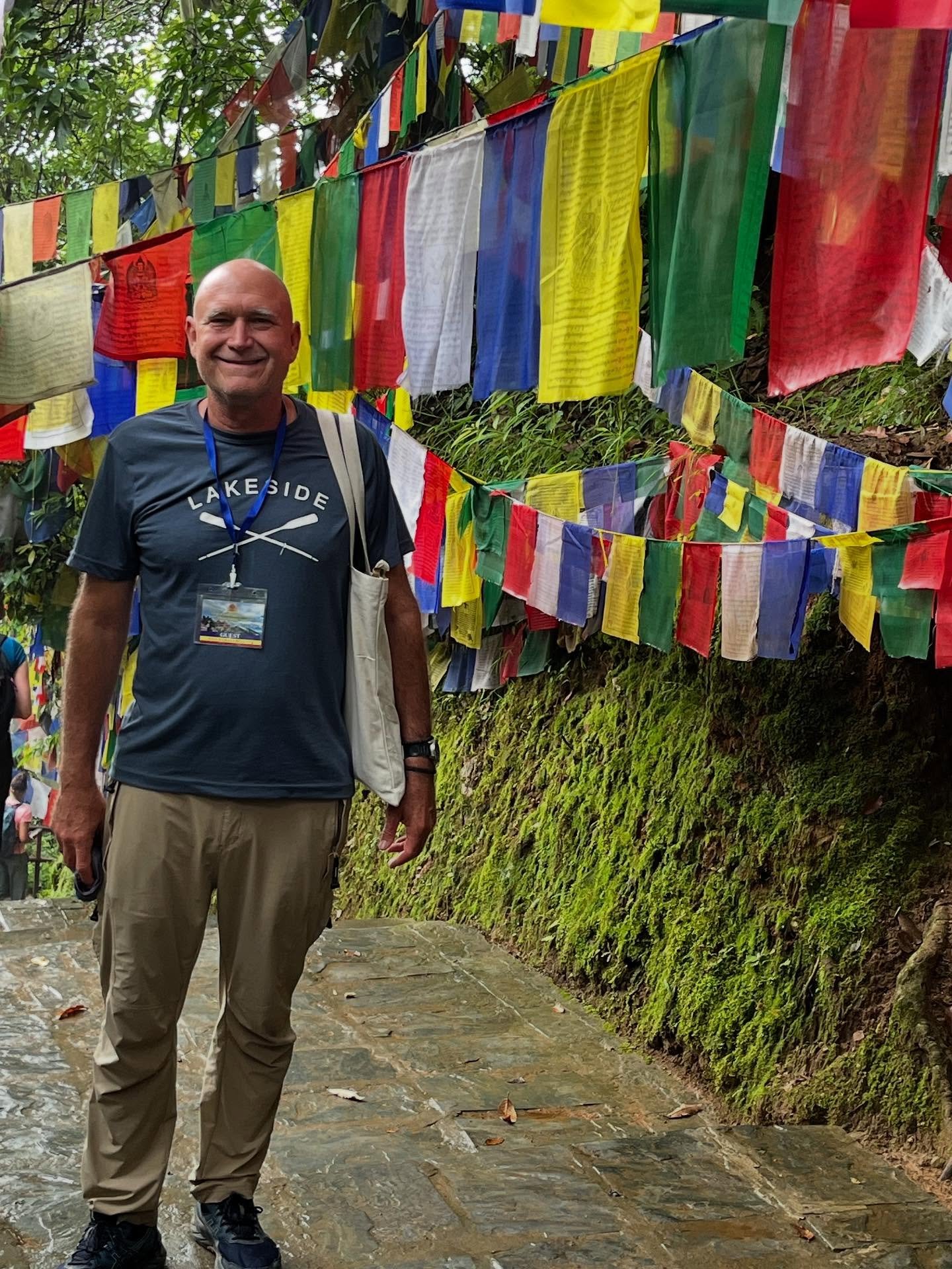 A smiling man wearing a dark blue t-shirt and beige pants standing on a stone path, with colorful prayer flags hanging behind him in a lush, green outdoor area.