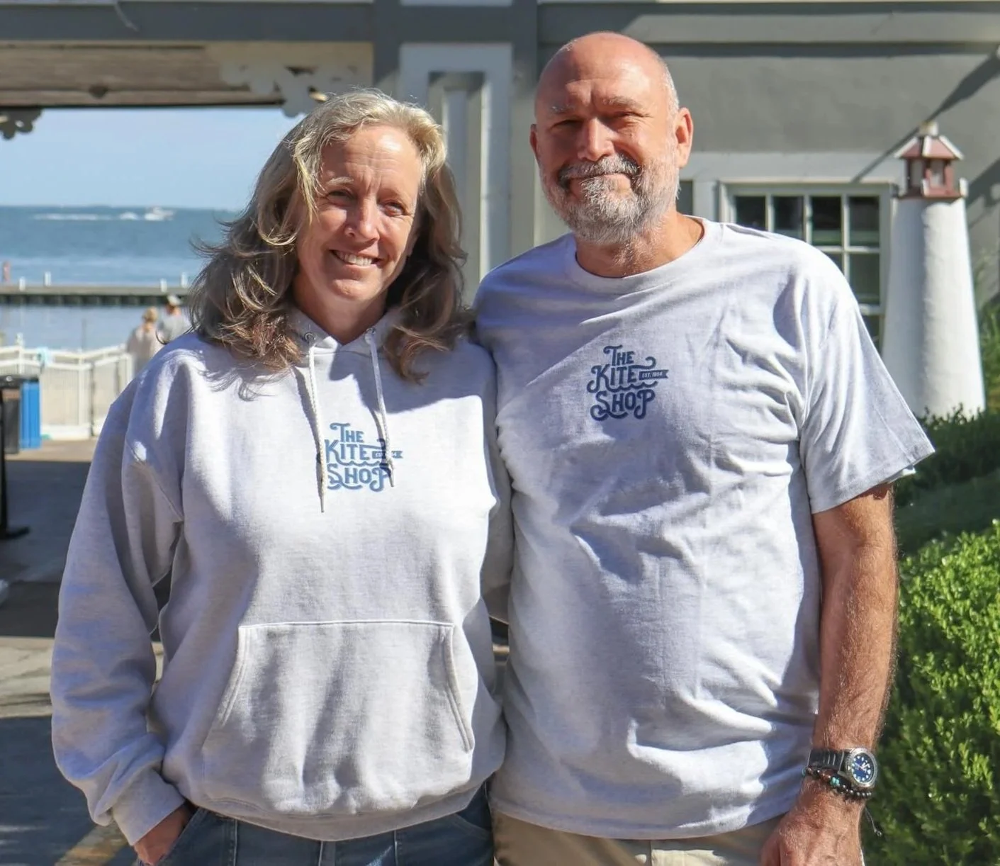 A smiling woman in a light gray hoodie and a smiling man in a light gray T-shirt, both wearing shirts with a logo for 'The Kite Shop' standing outdoors near the water with a pier and a lighthouse in the background.