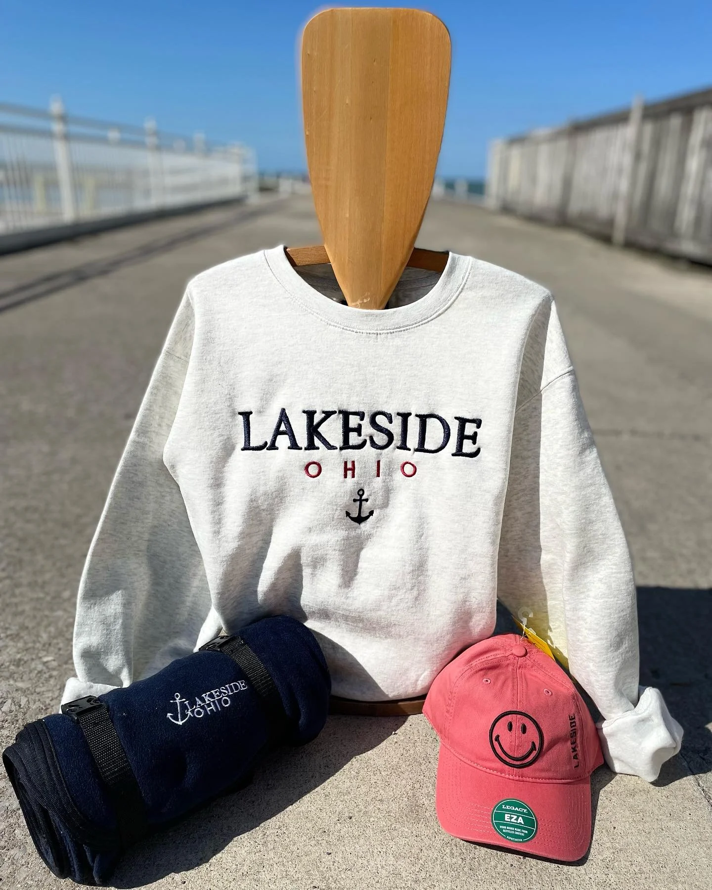 White sweatshirt with 'Lakeside Ohio' and an anchor embroidered on it, placed on a wooden hanger. There is a pink cap with a smiley face and 'Lakeside' written on the bill. A navy blue rolled-up blanket or towel with 'Lakeside Ohio' and an anchor embroidery next to the sweatshirt. The items are outdoors on a concrete surface with a walkway, fences, and a blue sky in the background.