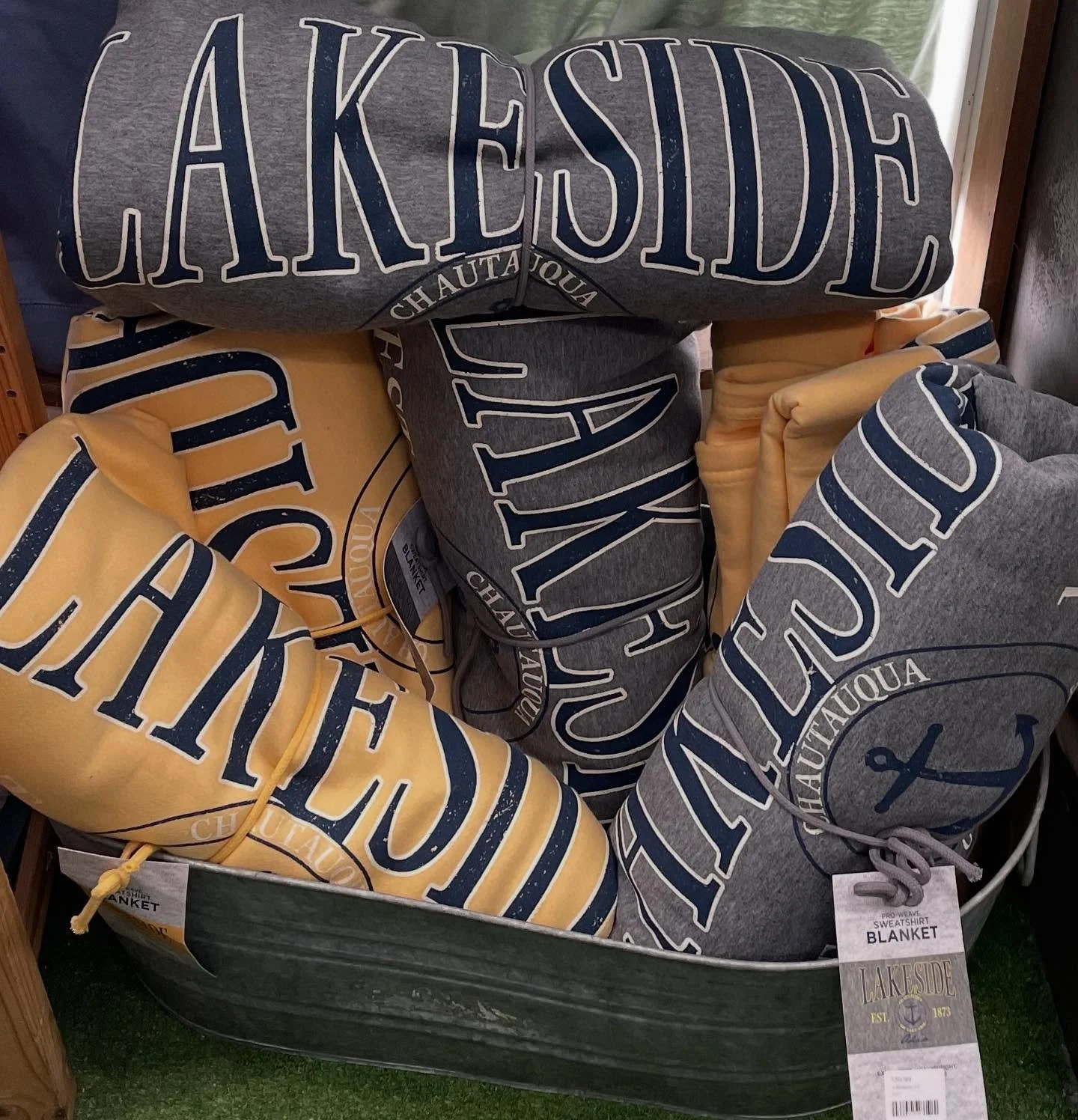 A display of folded sweatshirts or blankets in gray and yellow colors with nautical-themed text and designs, including an anchor, branding 'Lakeside,' and size tags.