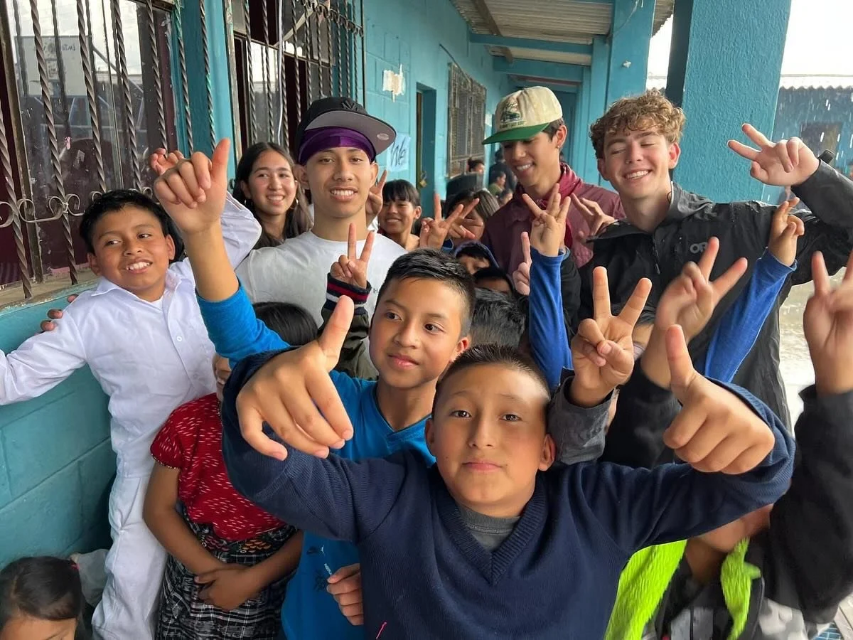 Group of children and teenagers smiling and making peace signs and thumbs-up gestures outdoors on a rainy day.