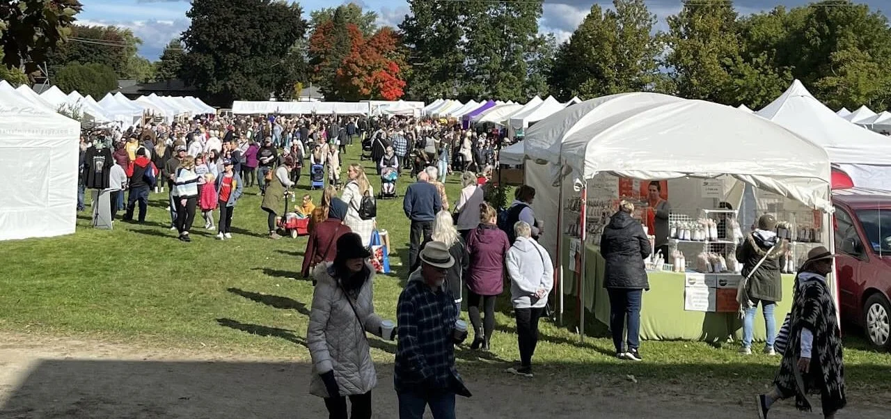 Crowd of people at an outdoor market with white tents, green grass, and colorful autumn trees in the background.