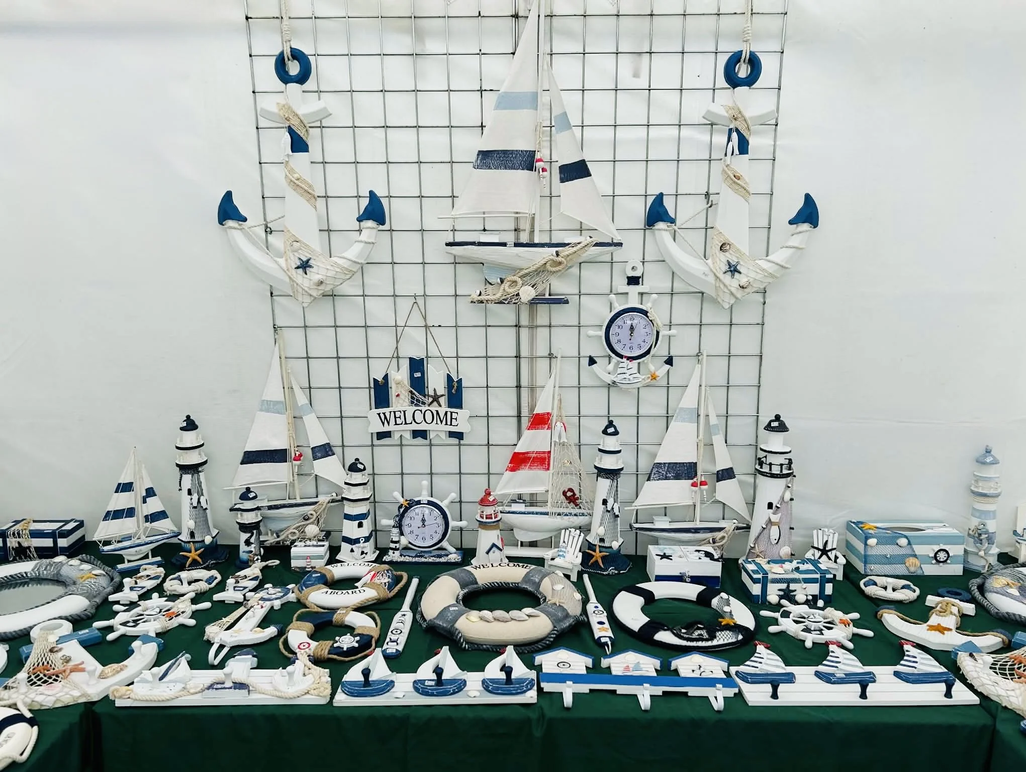 Display of nautical-themed decor items including anchors, sailboats, lighthouses, and clocks, arranged on a table with a green tablecloth and a wire grid backdrop.