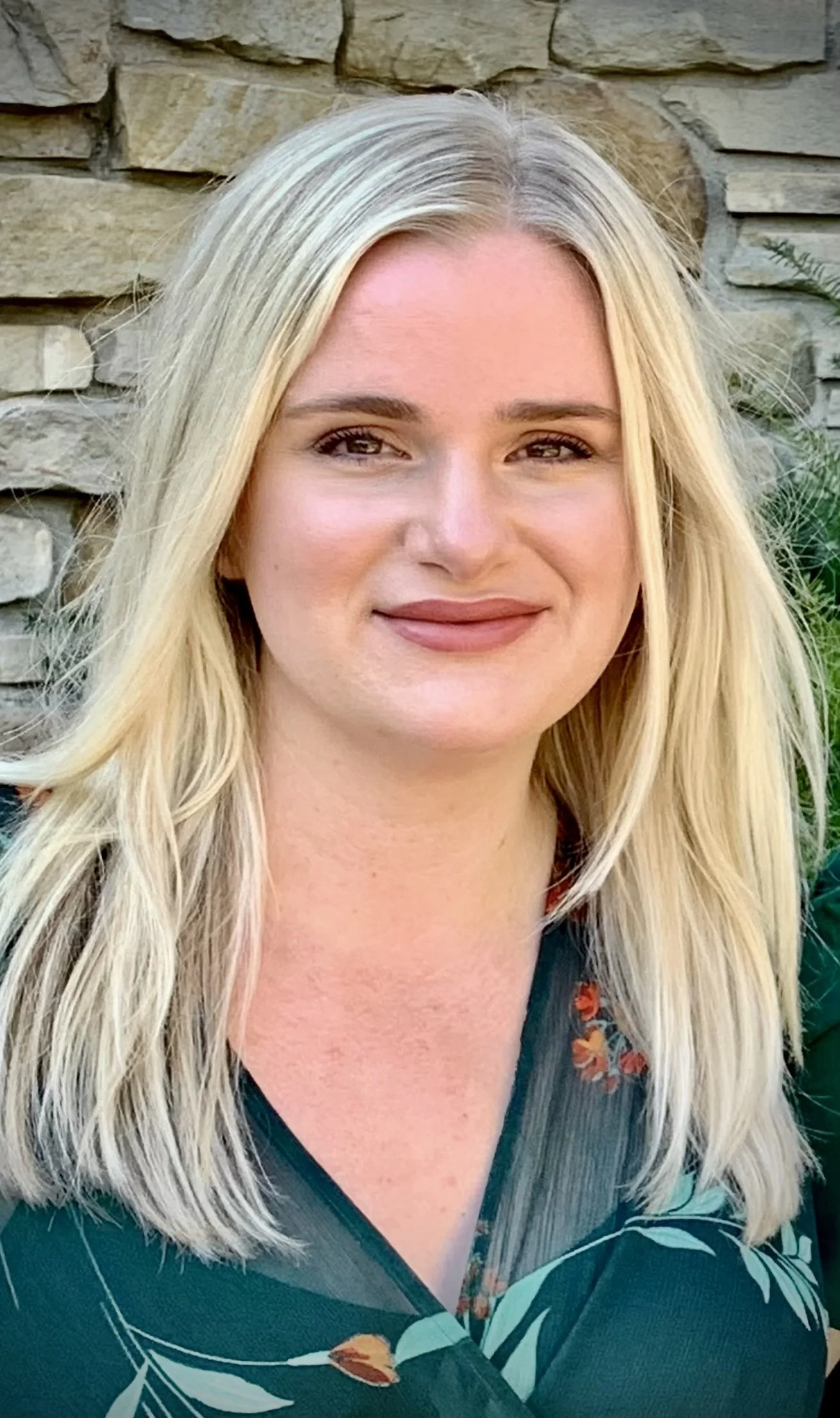 A woman with long blonde hair, light skin, and a gentle smile, standing outdoors in front of a stone wall and some green foliage, wearing a dark green top with a floral pattern.