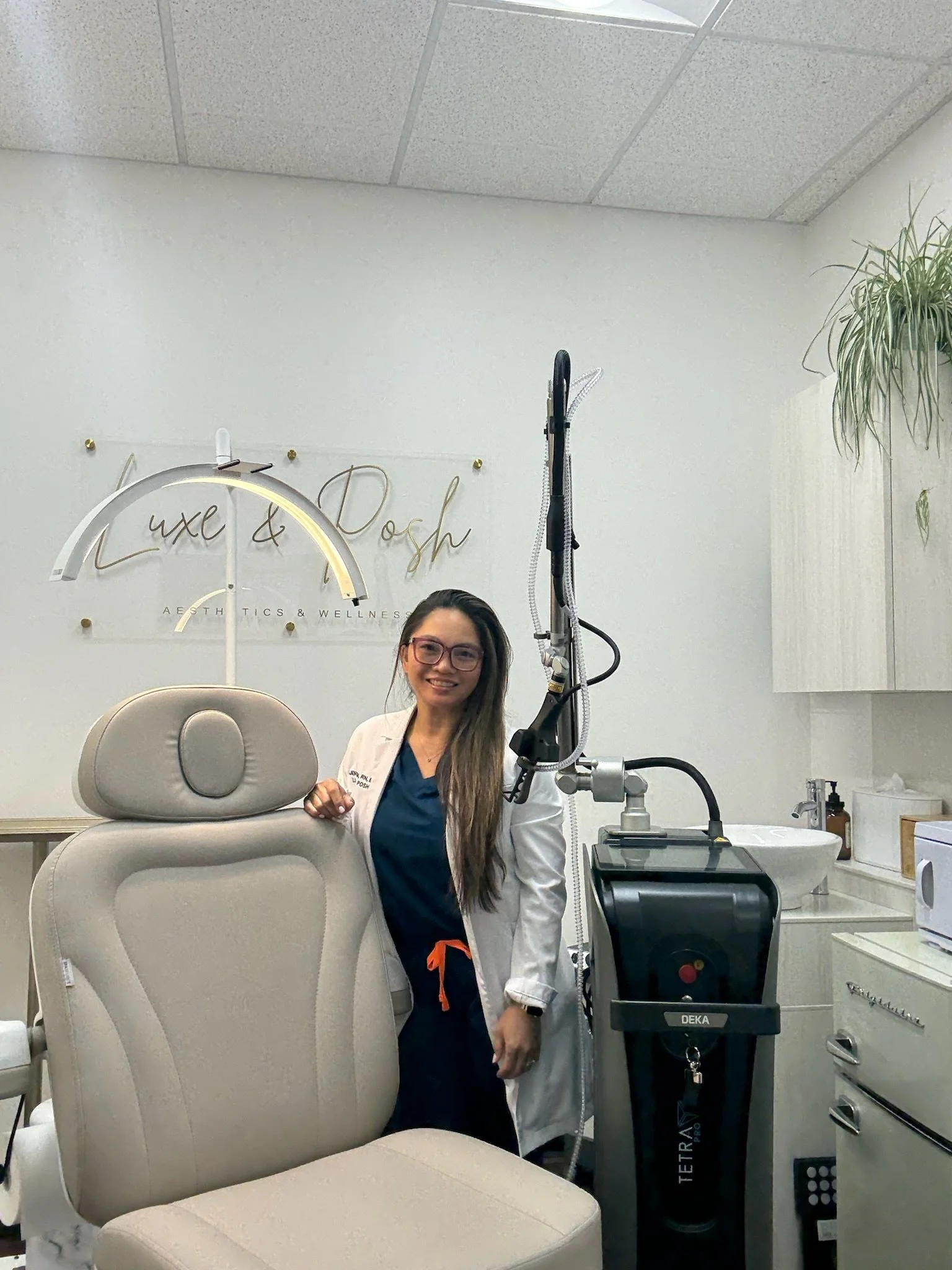 A smiling woman with long hair and glasses standing next to a medical treatment chair in a wellness clinic, with medical equipment and a sign reading 'Luxe & Dosh Aesthetics & Wellness' in the background.