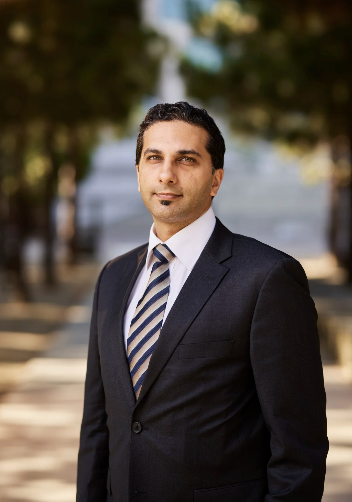 A man in a black suit, white shirt, and striped tie standing outdoors with trees in the background.