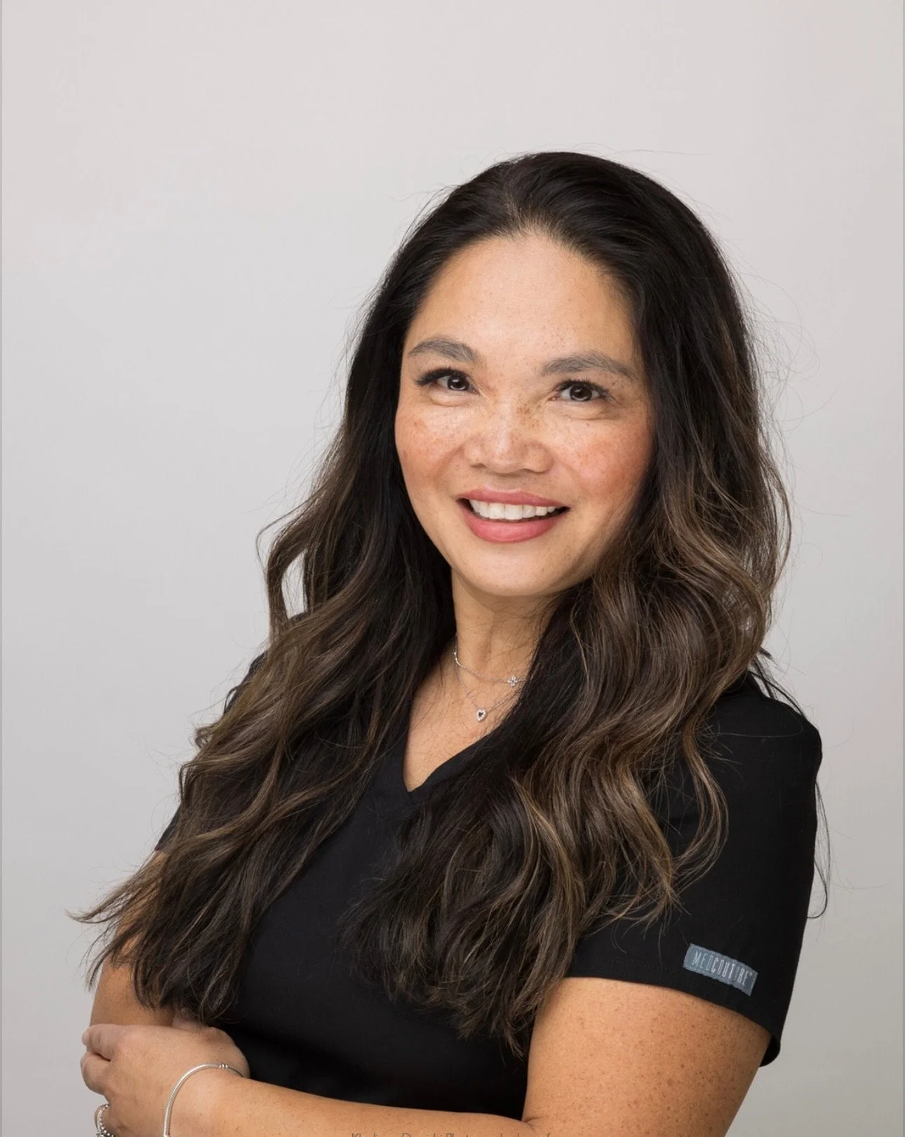 Woman with long, wavy brown hair wearing a black shirt and silver jewelry against a light gray background.