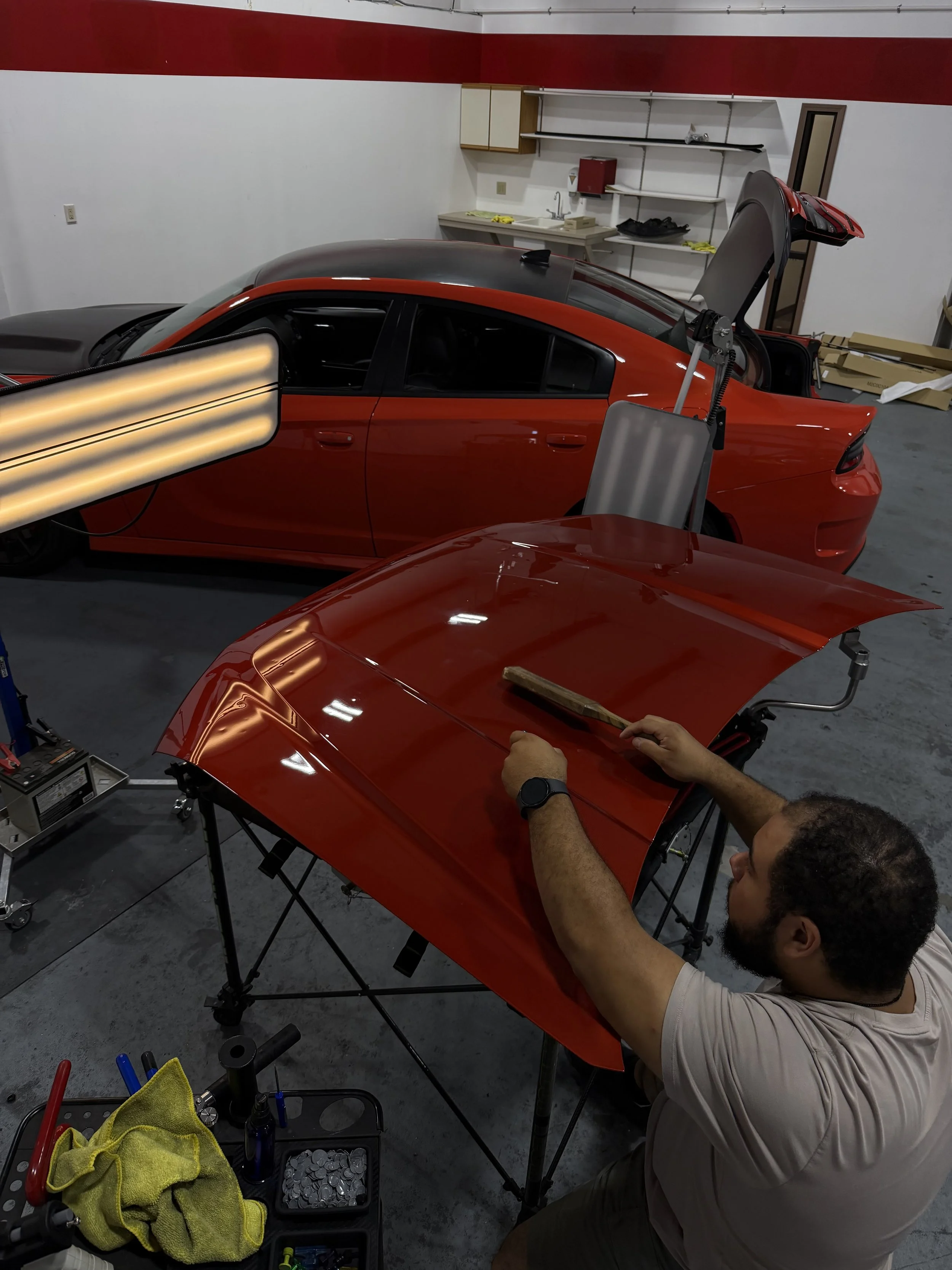 Person working on assembling a red sports car in a garage or workshop, with tools and parts on a work table and shelves in the background.