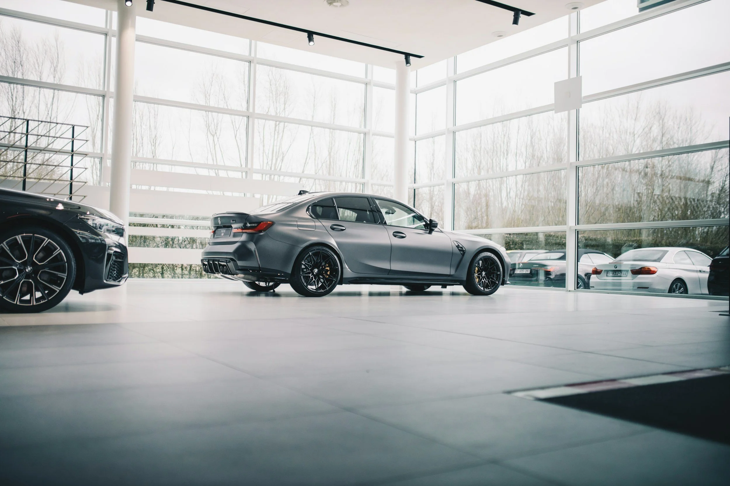 Inside a modern car dealership showroom with large glass windows, displaying a sleek gray sports car and parts of two other cars.