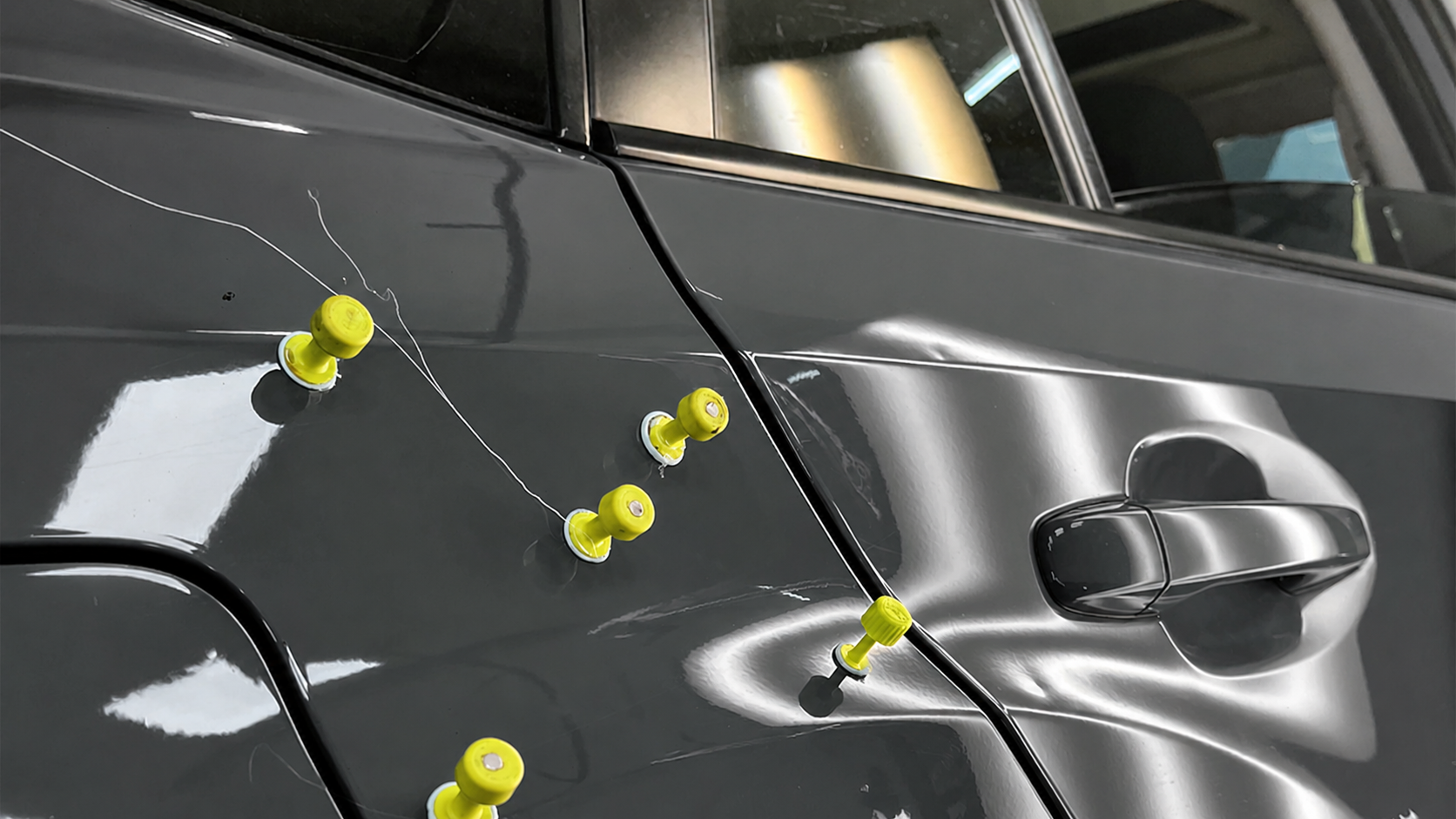 Close-up of dark gray car door with yellow push pins and strings attached, indicating a repair or inspection process in an automotive workshop.