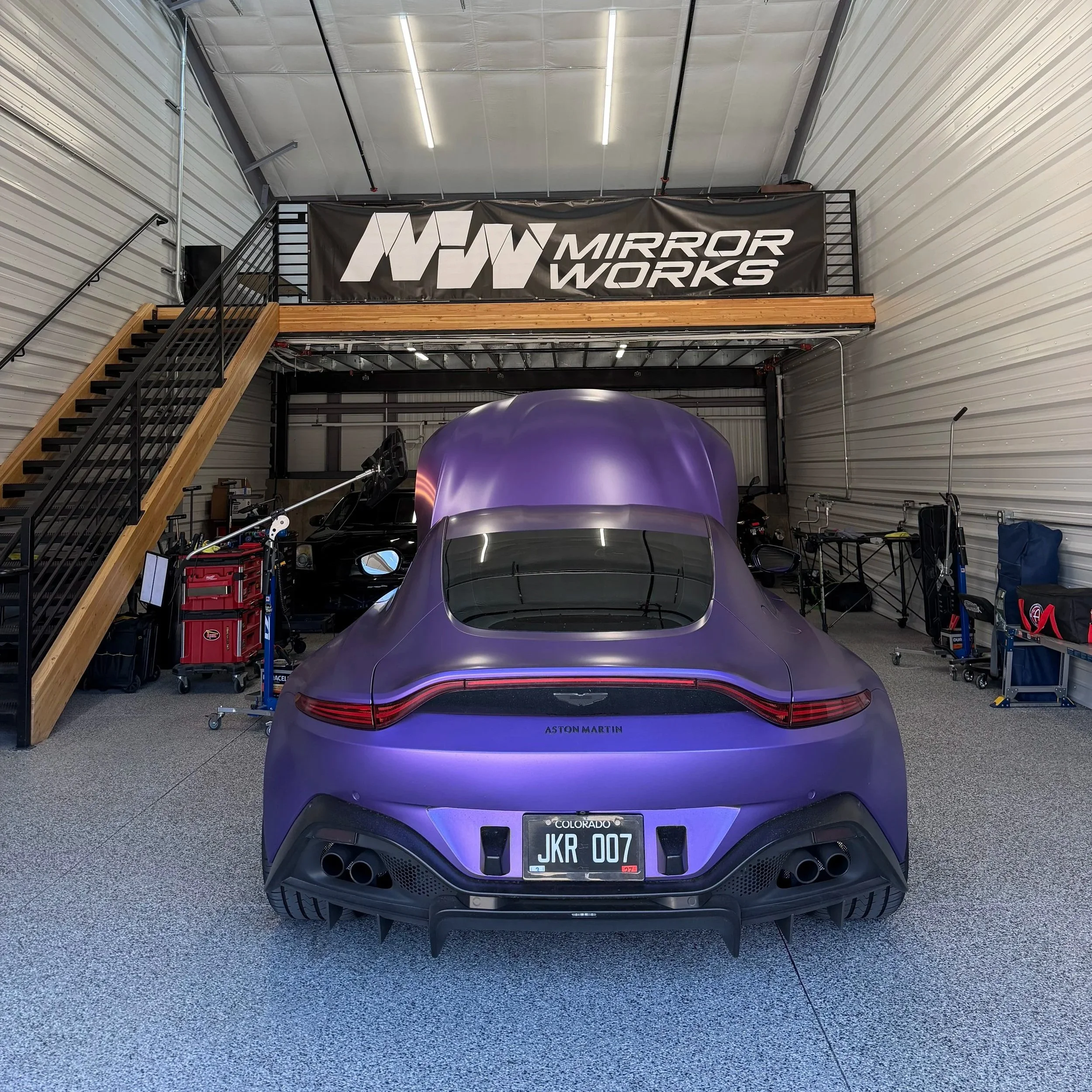 A purple Aston Martin car parked inside a spacious industrial garage with metal walls. There is a wooden staircase with black railings leading to a second level with a large black banner reading 'WW MIRROR WORKS'. The garage contains various tools and equipment.